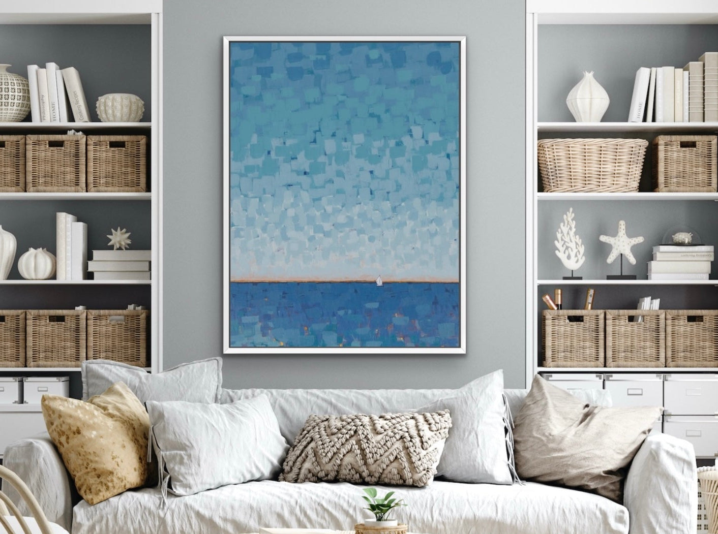 A framed vertical art print featuring a textured blue and white ocean scene hangs on a gray wall above a white sofa in a styled living room with shelving.