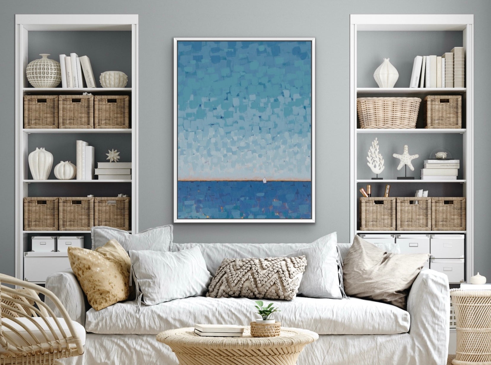 A framed vertical art print featuring a textured blue and white ocean scene hangs on a gray wall above a white sofa in a styled living room with shelving.