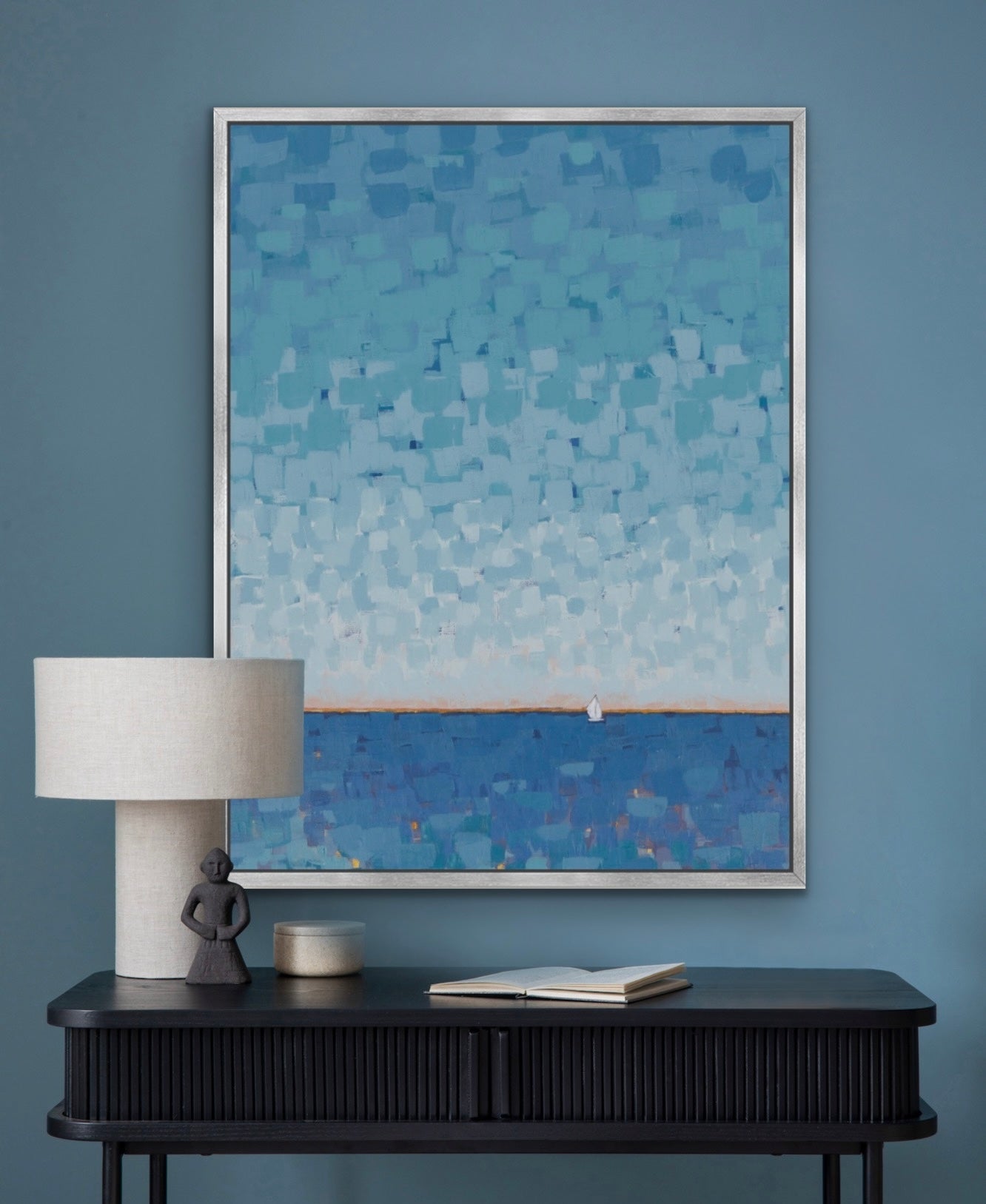 A framed abstract painting featuring a blue and white textured pattern of water and sky, hung on a blue wall above a dark console table with a lamp and books.
