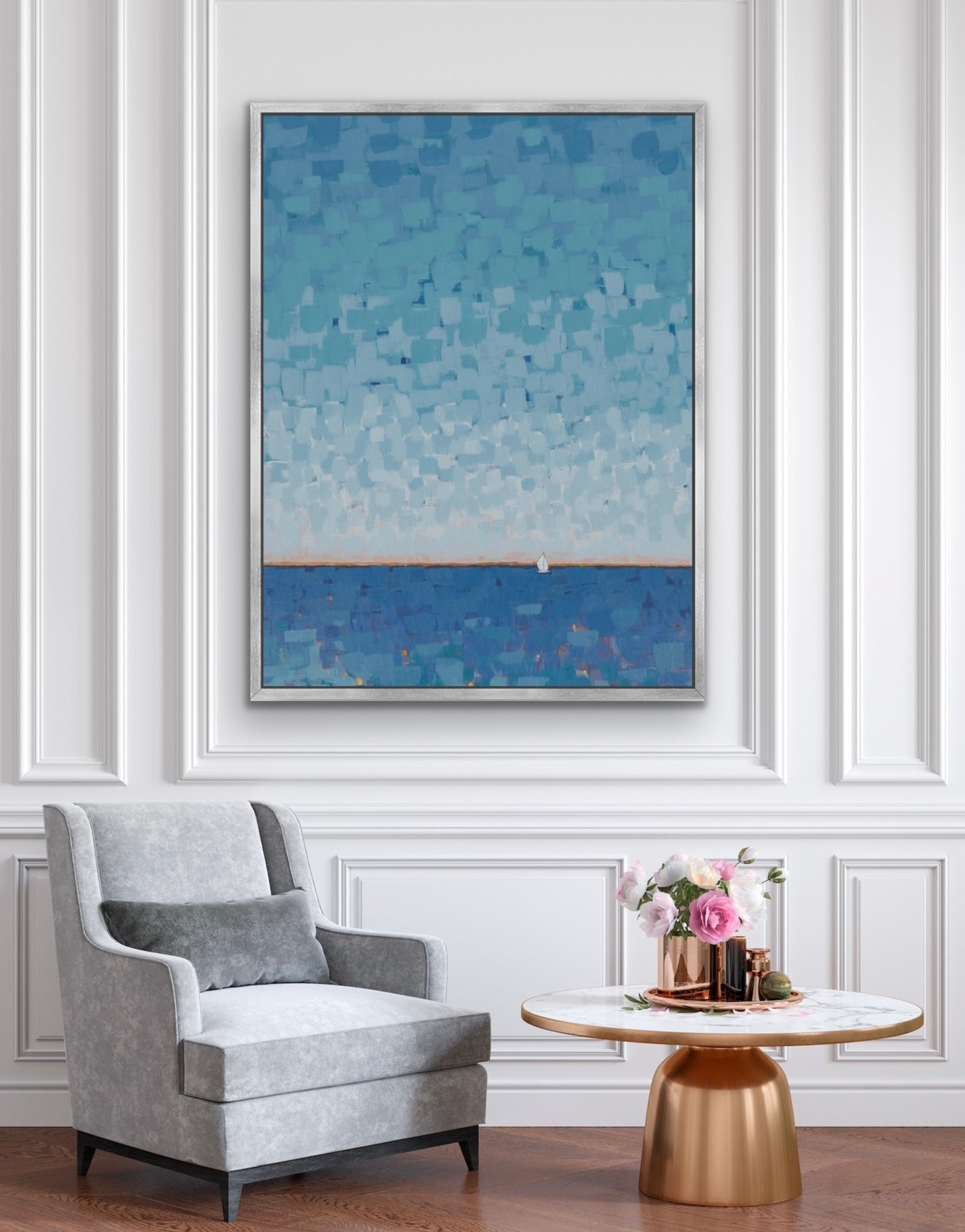 A framed abstract landscape painting featuring a blue sky and sea, displayed on a white paneled wall above a gray armchair and a small round table with flowers.