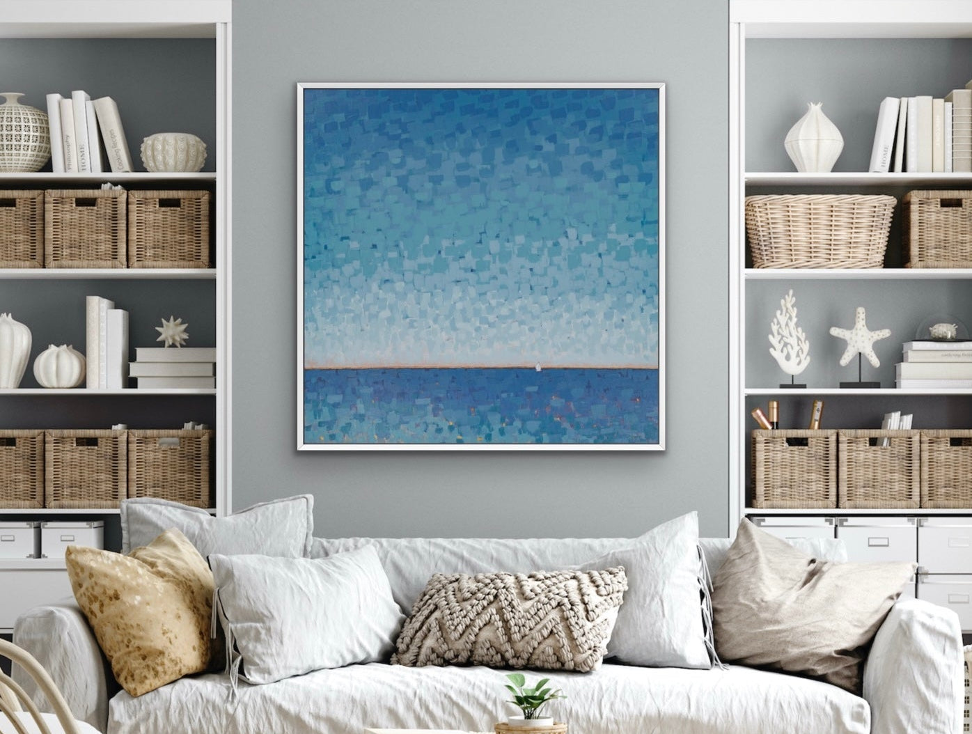 A framed square canvas print of a blue textured seascape hangs on a gray wall above a white sofa in a styled living room with shelving and decorative items.
