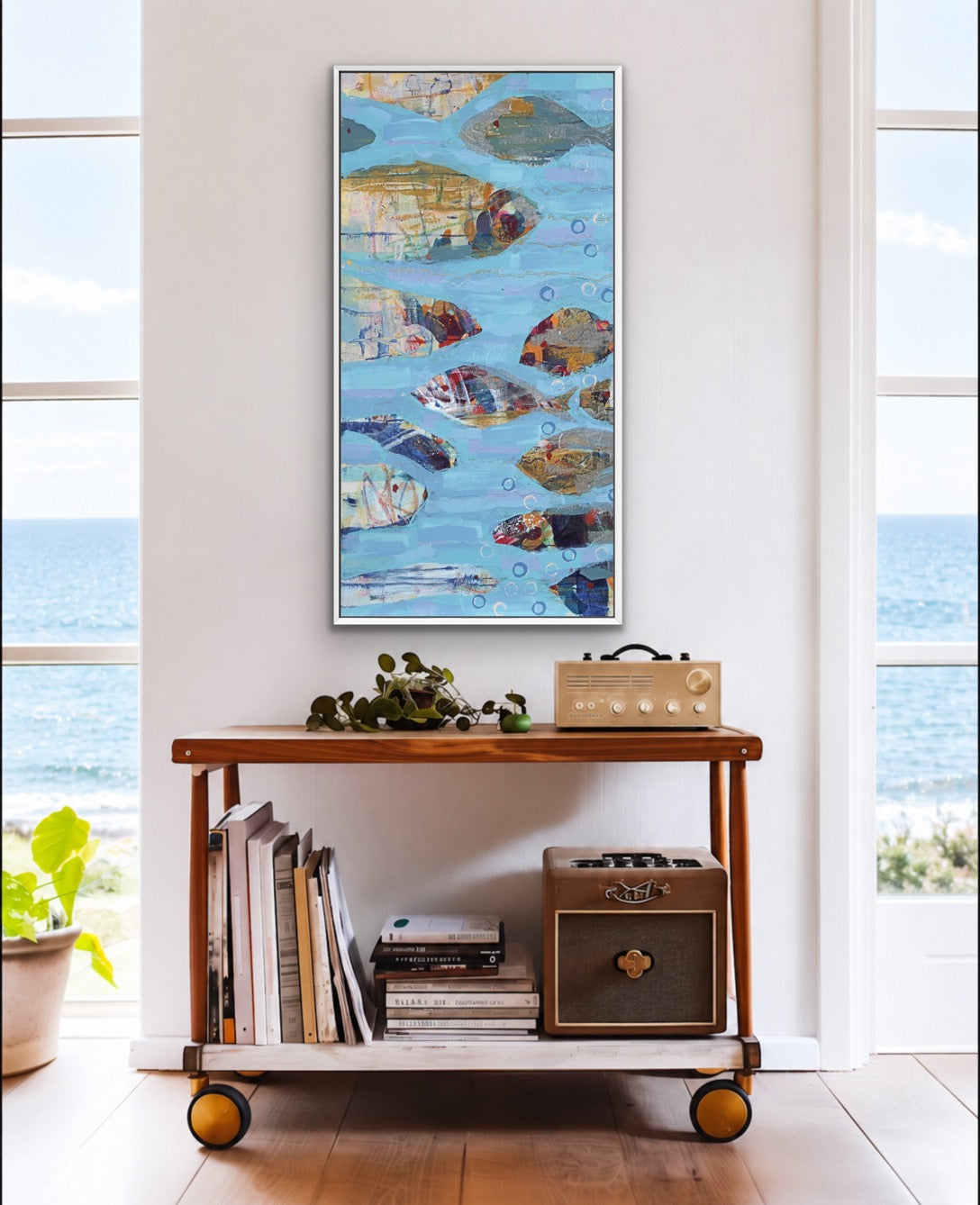 A framed vertical art print depicting colorful painted fish swimming in a blue water scene, displayed on a wooden rolling cart in a bright room with an ocean view background.