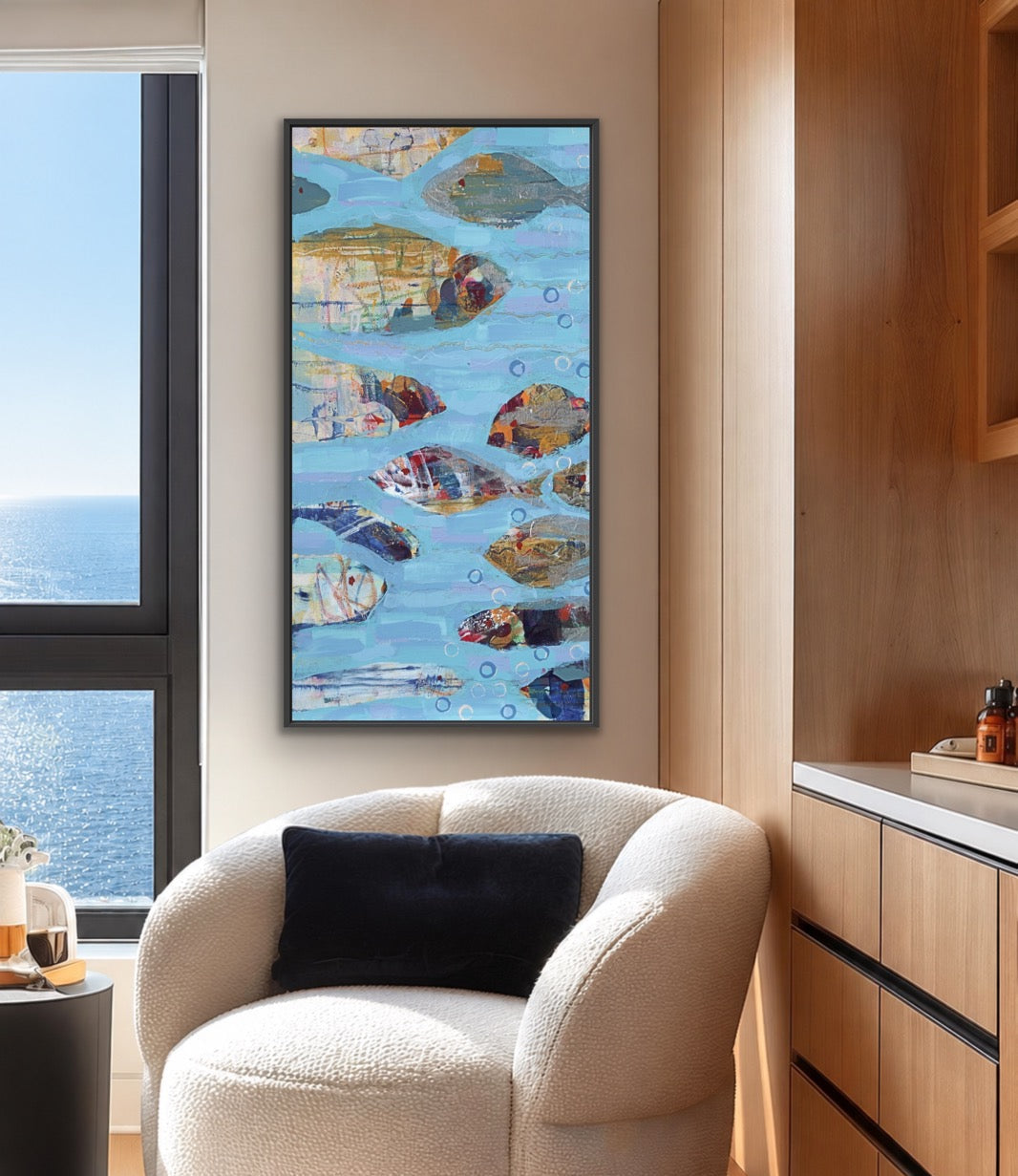 A framed vertical canvas painting of colorful painted fish on a blue water background, displayed on a beige wall in a modern living room setting above a cream boucle armchair.