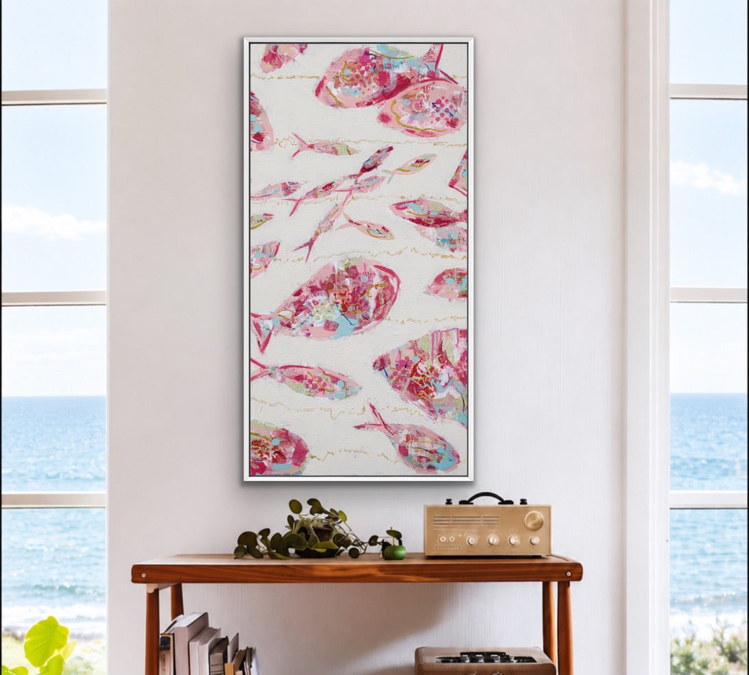 A vertical framed art print featuring pink and red fish illustrations on a white background, styled on a wooden rolling cart in a bright room with an ocean view.
