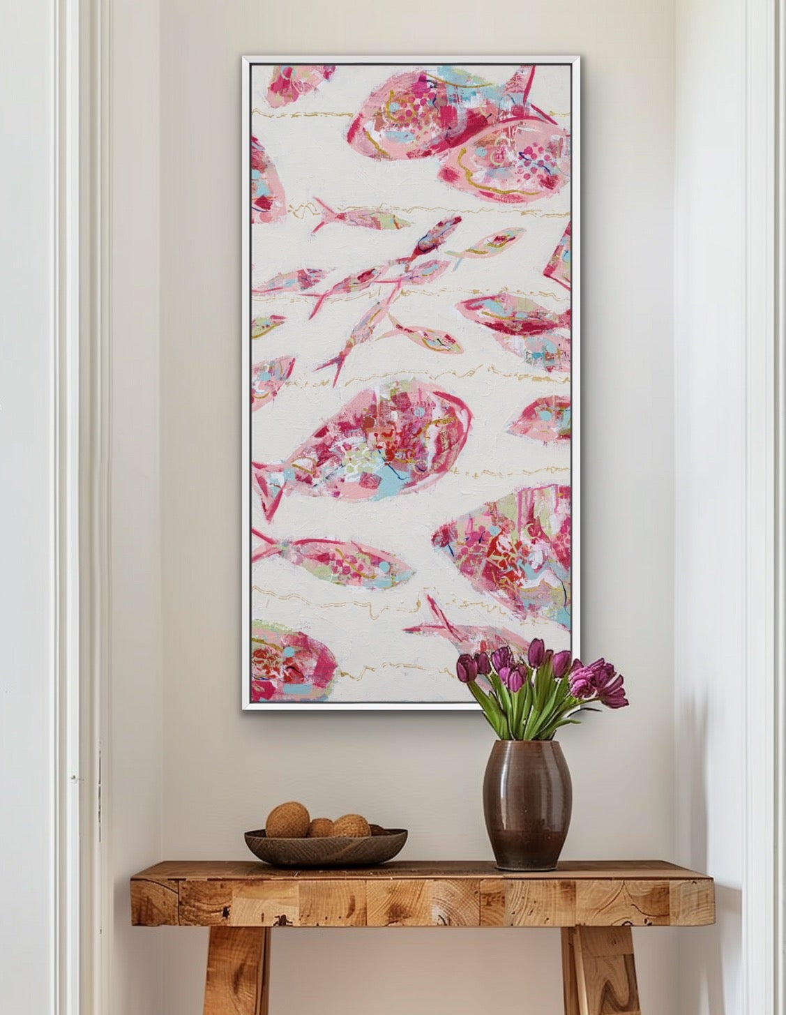 A framed vertical wall art print featuring a watercolor pattern of pink, red, and blue fish on a white background, styled in a home entryway above a rustic wooden console table with a vase of purple tulips.