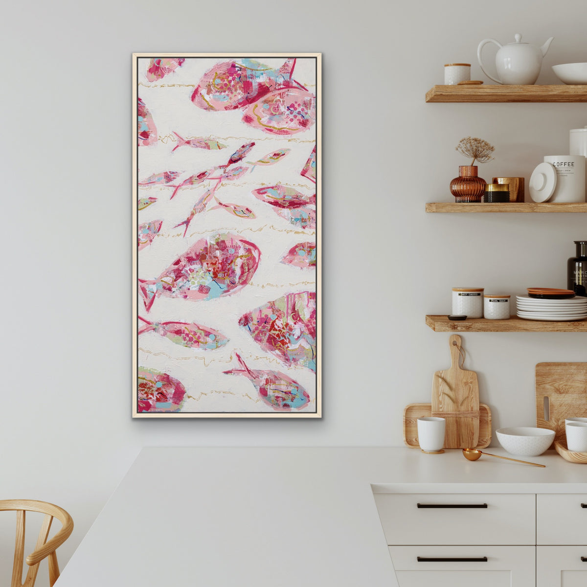 A vertical framed wall art print featuring a white background with pink, red, and blue watercolor fish illustrations, displayed in a modern kitchen setting.