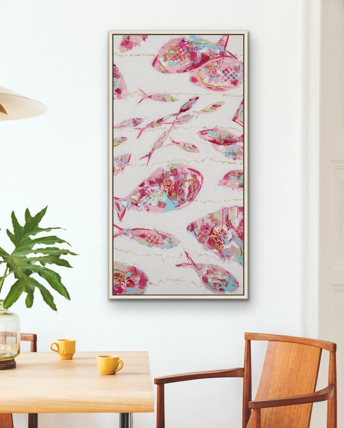 A vertical framed wall art print featuring a pink and blue fish pattern on a white background, styled in a modern dining room setting above a wooden table and chairs.