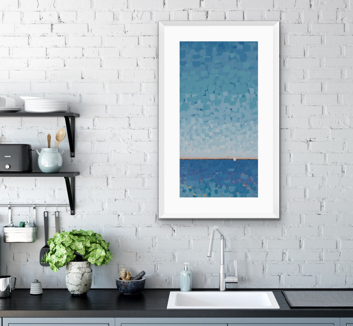 A framed vertical art print depicting a blue textured seascape with a small sailboat, displayed on a white brick wall above a kitchen sink.