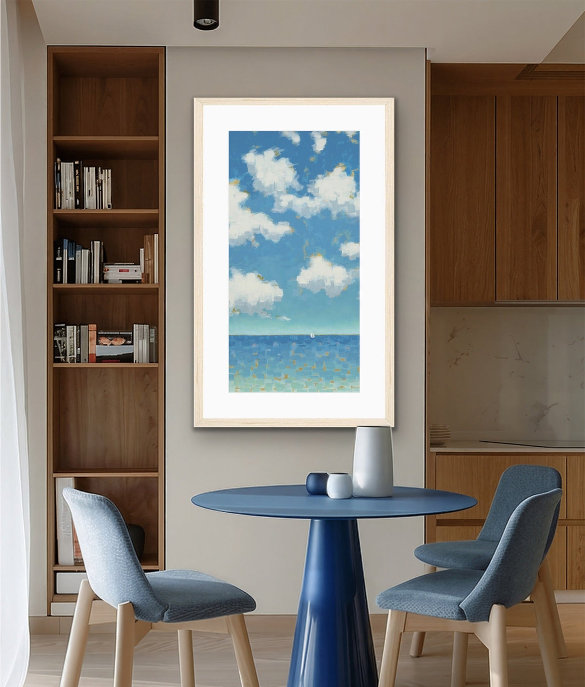 A framed vertical art print depicting a blue sky with white clouds over a sea, displayed on a beige wall in a modern dining room setting with a blue table and chairs.