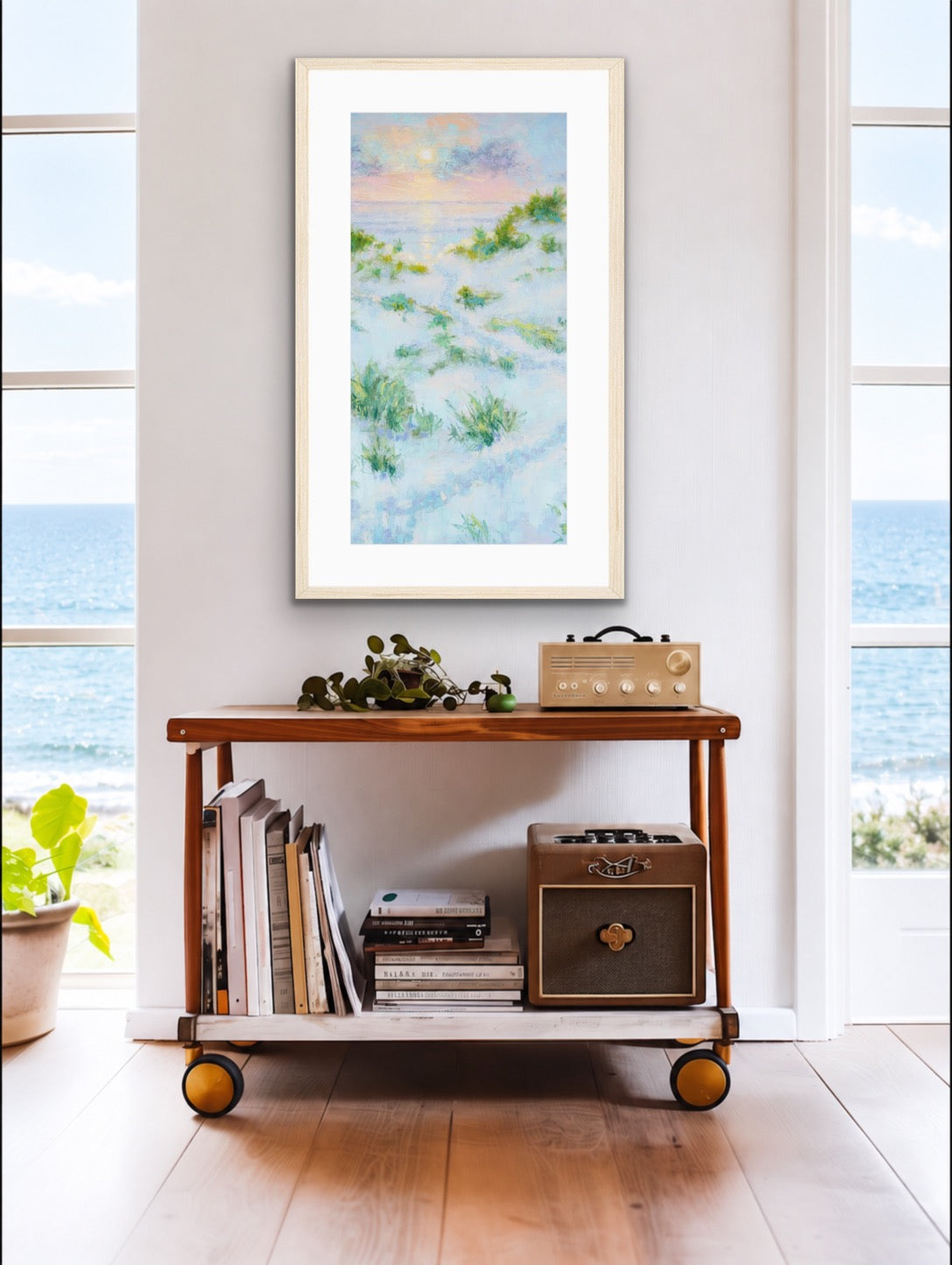 A framed abstract coastal landscape print hangs on a white wall above a wooden rolling cart in a bright room with windows overlooking the ocean. The artwork shows soft blue, white, and green tones with a pale sky at the top.