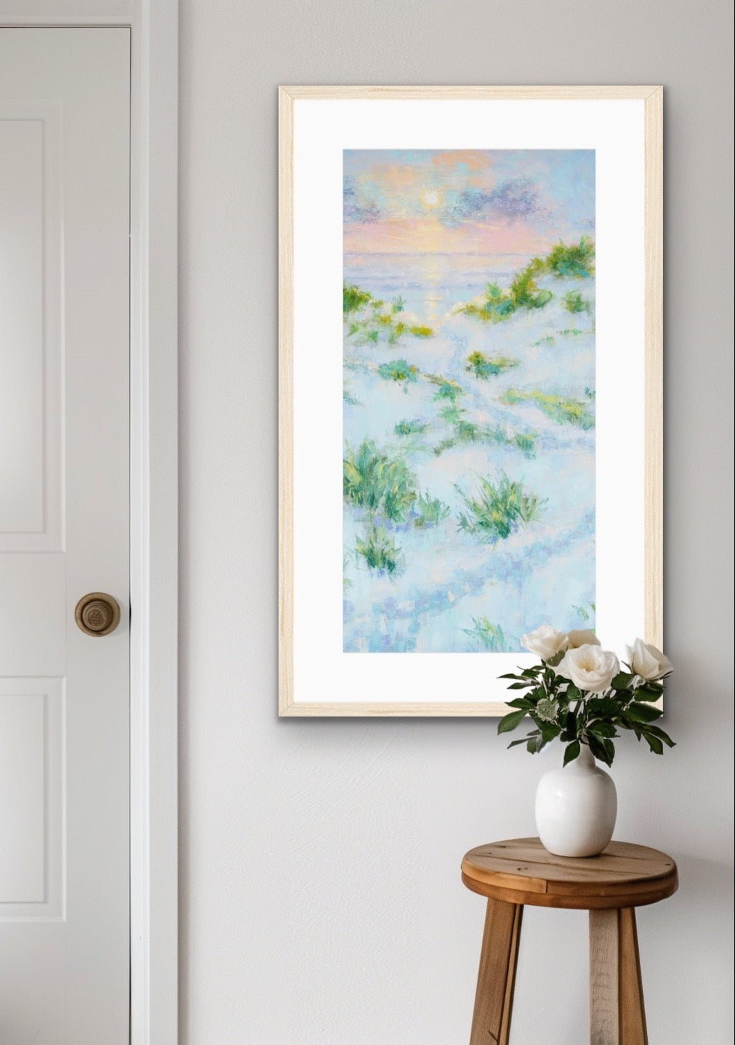 A framed vertical landscape painting of grassy dunes under a pastel sky, displayed on a light gray wall in a styled room next to a white door and a small wooden stool with a vase of white flowers.