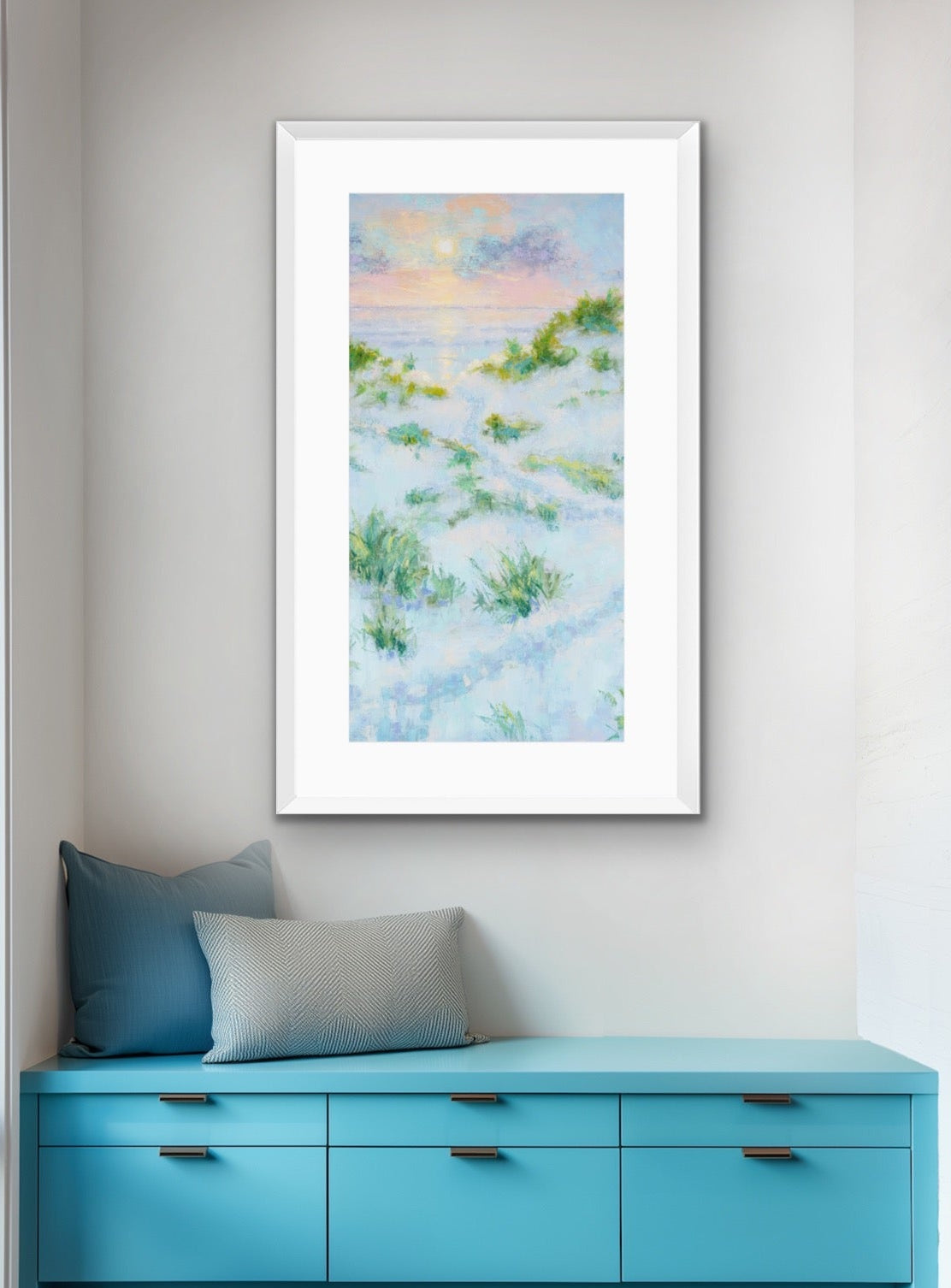 A framed vertical art print depicting a soft, impressionistic beach or dune landscape in pastel blue, green, and pink tones, displayed on a light gray wall above a bright turquoise bench with pillows.