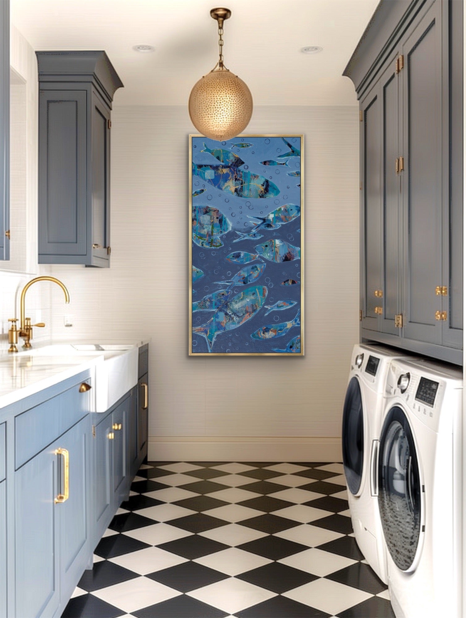 A framed vertical wall art print featuring a blue fish school pattern hangs on a light gray wall in a modern laundry room with gray cabinets, a black-and-white checkered floor, and a gold pendant light.