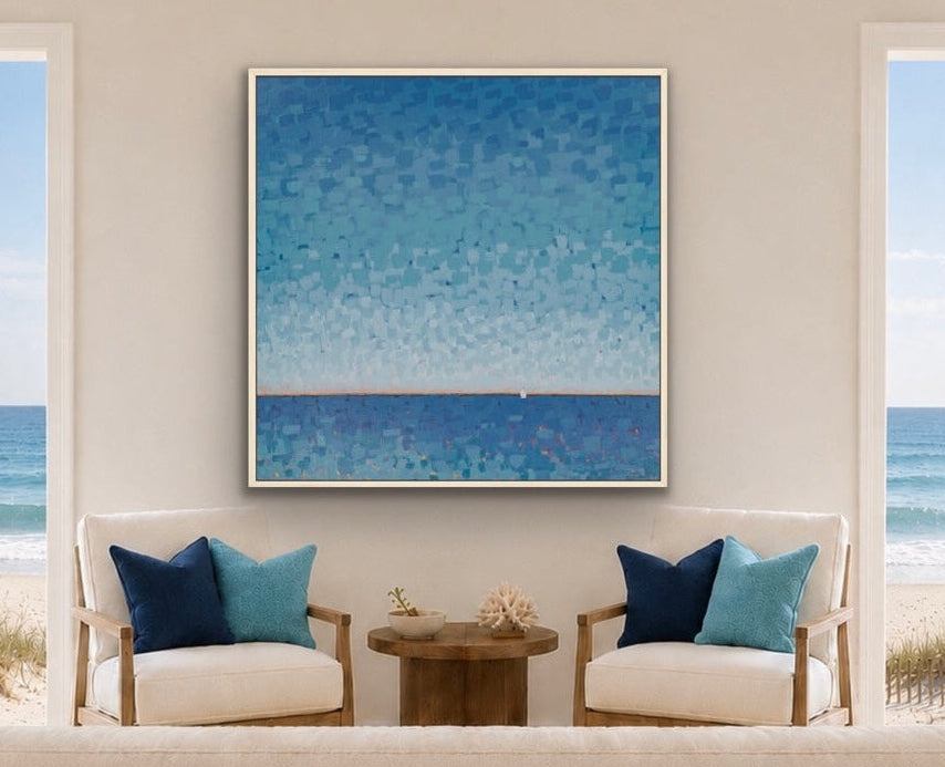 A square framed abstract blue painting hangs on a beige wall in a bright living room with two white armchairs, blue and teal pillows, a wooden coffee table, and large windows overlooking the ocean and beach.