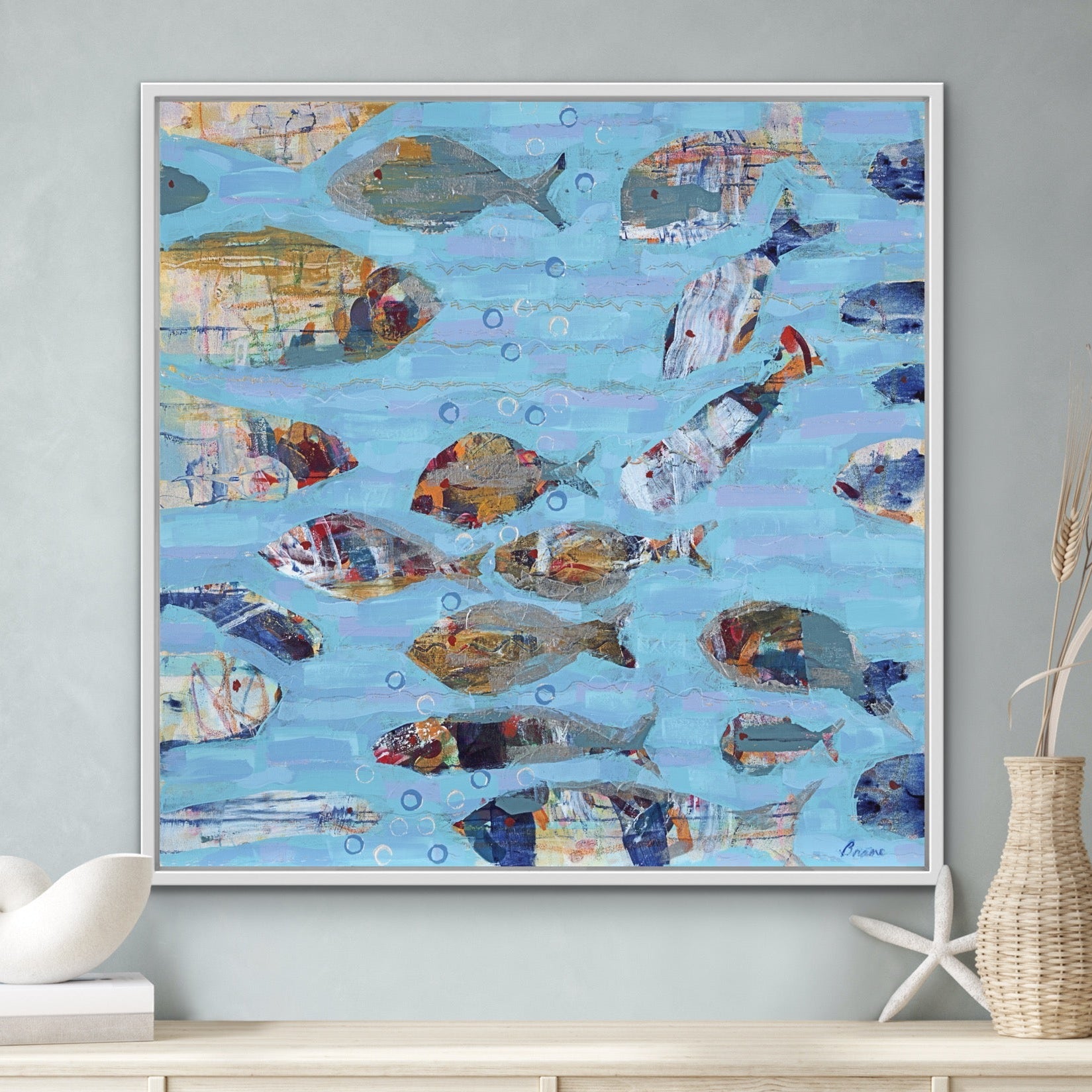 Beach house wall art with expressive fish forms and aqua ocean tones displayed on a wall above a wooden cabinet.