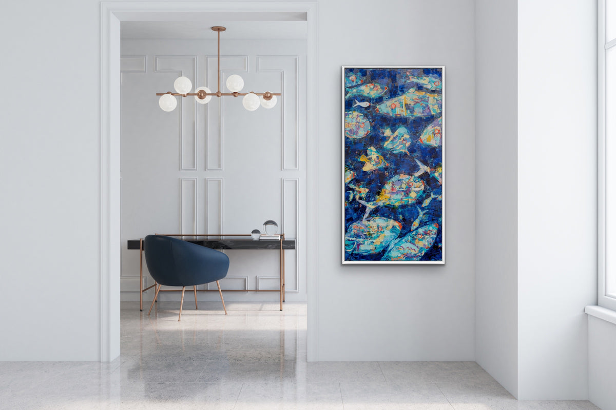 A vertical framed abstract painting featuring blue, teal, and gold brushstrokes, displayed on a white wall in a modern, bright interior room with a desk and chair visible in the background.