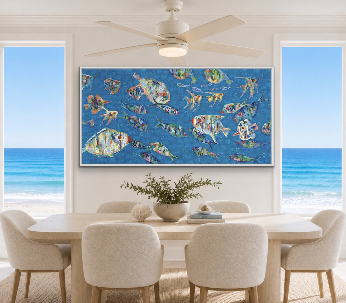 A large framed wall art print depicting colorful tropical fish on a bright blue background, displayed above a dining table in a modern, sunlit room with ocean views through tall windows.