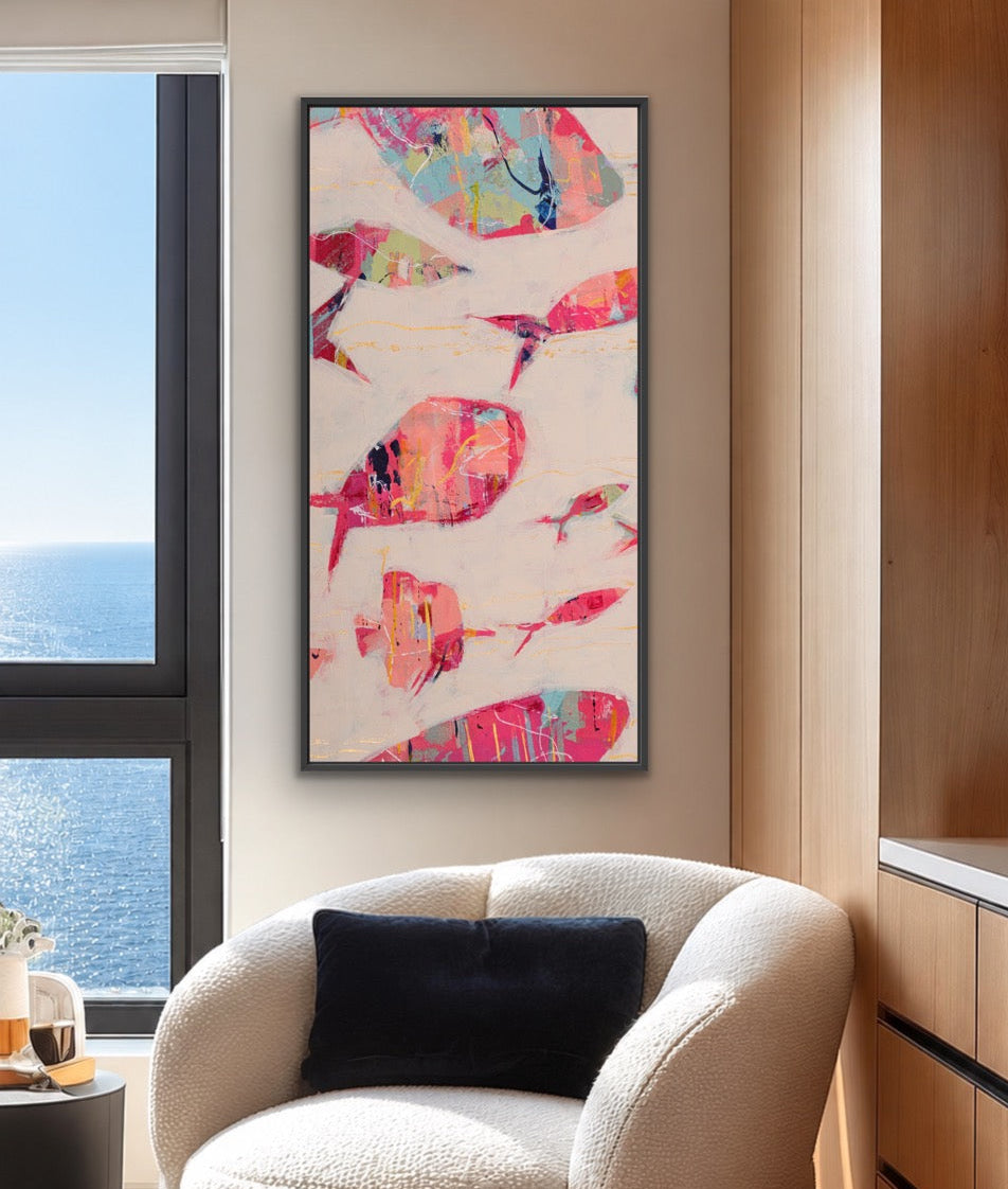 A framed abstract wall art print featuring colorful painted fish on a light background, displayed in a modern living room setting above a cream-colored armchair.