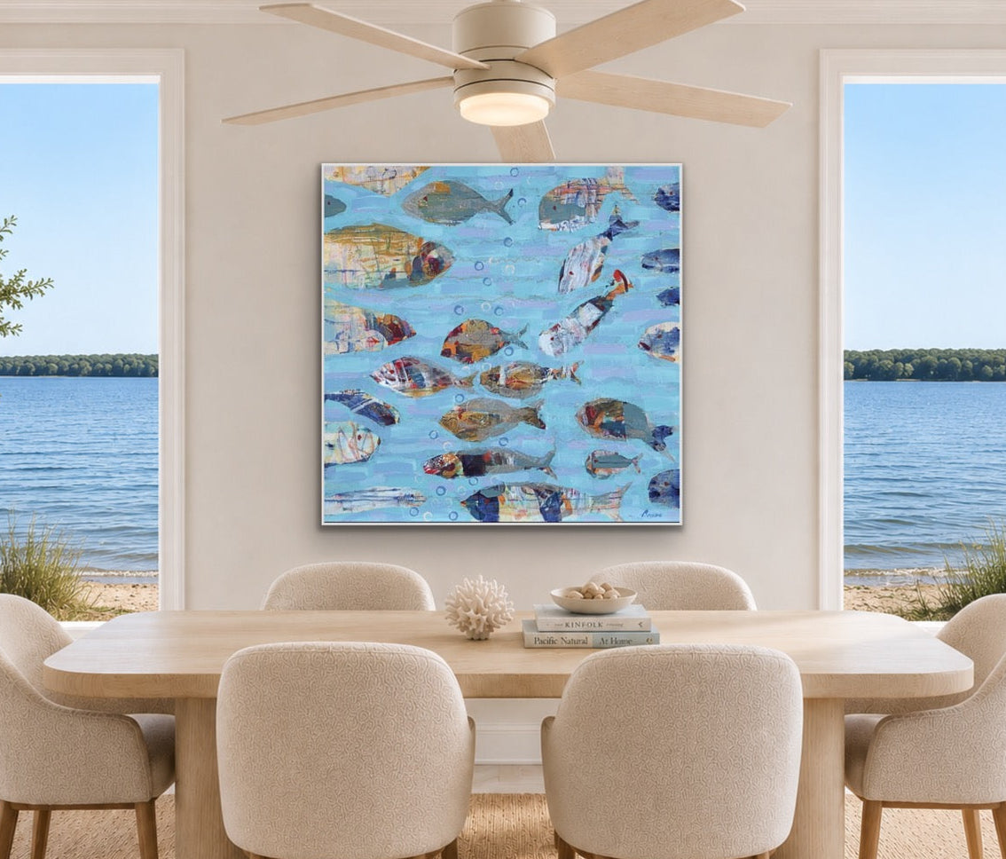 A square canvas wall art print depicting a school of colorful fish swimming on a light blue water background, displayed in a modern dining room setting with a wooden dining table and six upholstered chairs, with large windows showing a lake view.