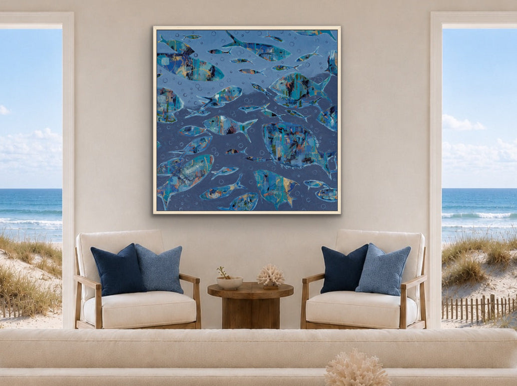 A framed abstract blue fish painting hangs on a beige wall in a bright, modern living room with a white sofa, wooden coffee table, and large windows overlooking the ocean and beach dunes.