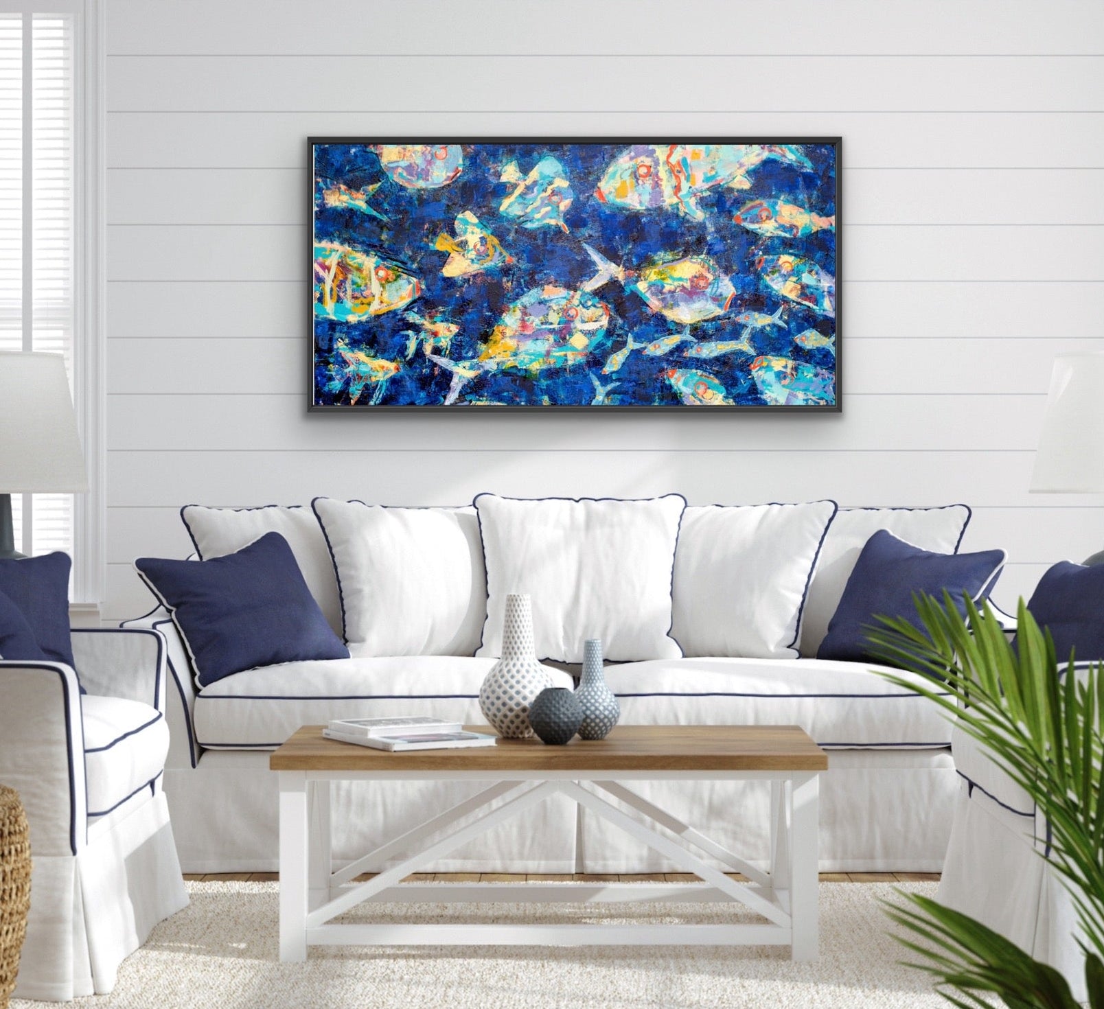 A framed abstract canvas print with vibrant blue, gold, and white brushstrokes hangs on a white shiplap wall above a white sofa with navy blue pillows in a bright, modern living room.