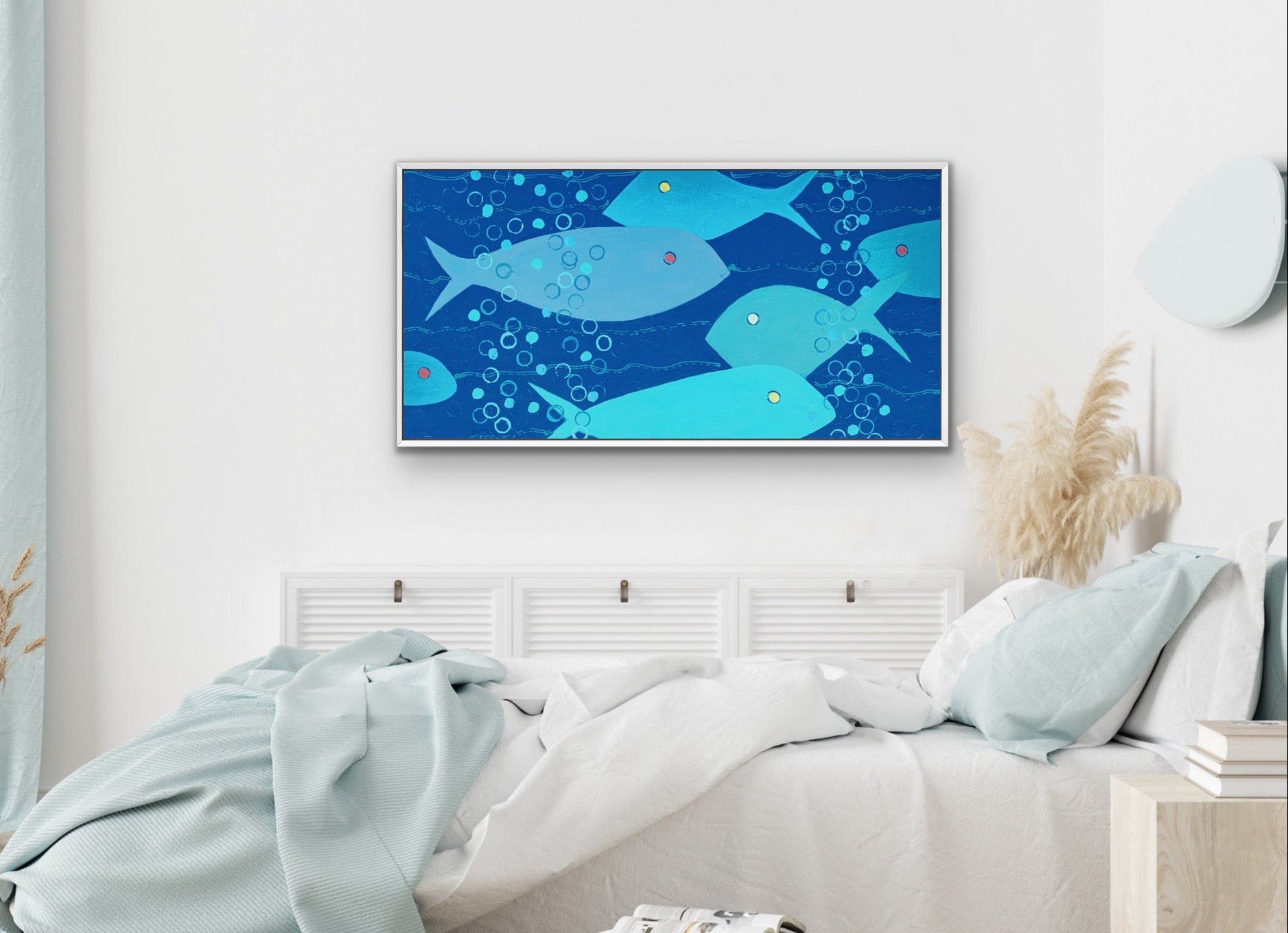 A framed wall art print featuring a blue underwater scene with stylized fish and bubbles, displayed above a neatly made bed in a bright, modern bedroom.