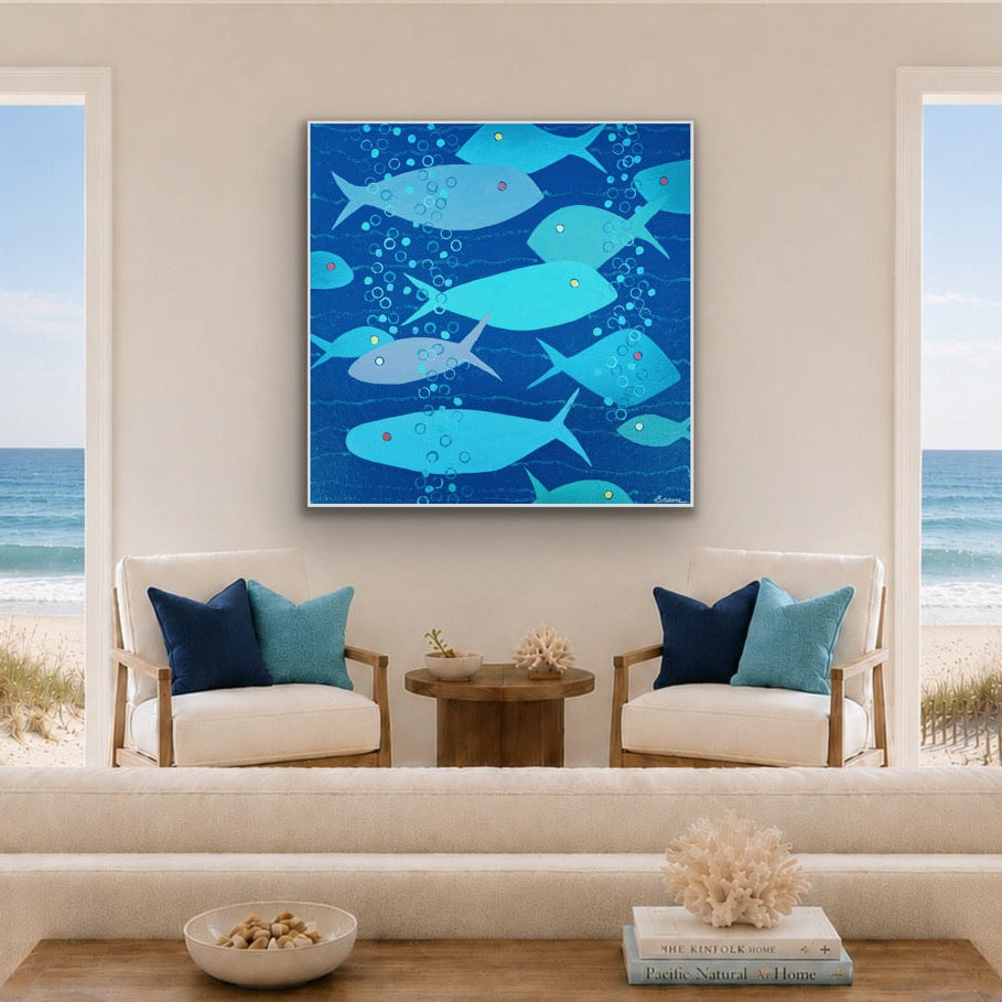 A square framed wall art print featuring a school of blue fish on a deep blue background, displayed above a white sofa in a bright, modern living room with beach views through large windows.