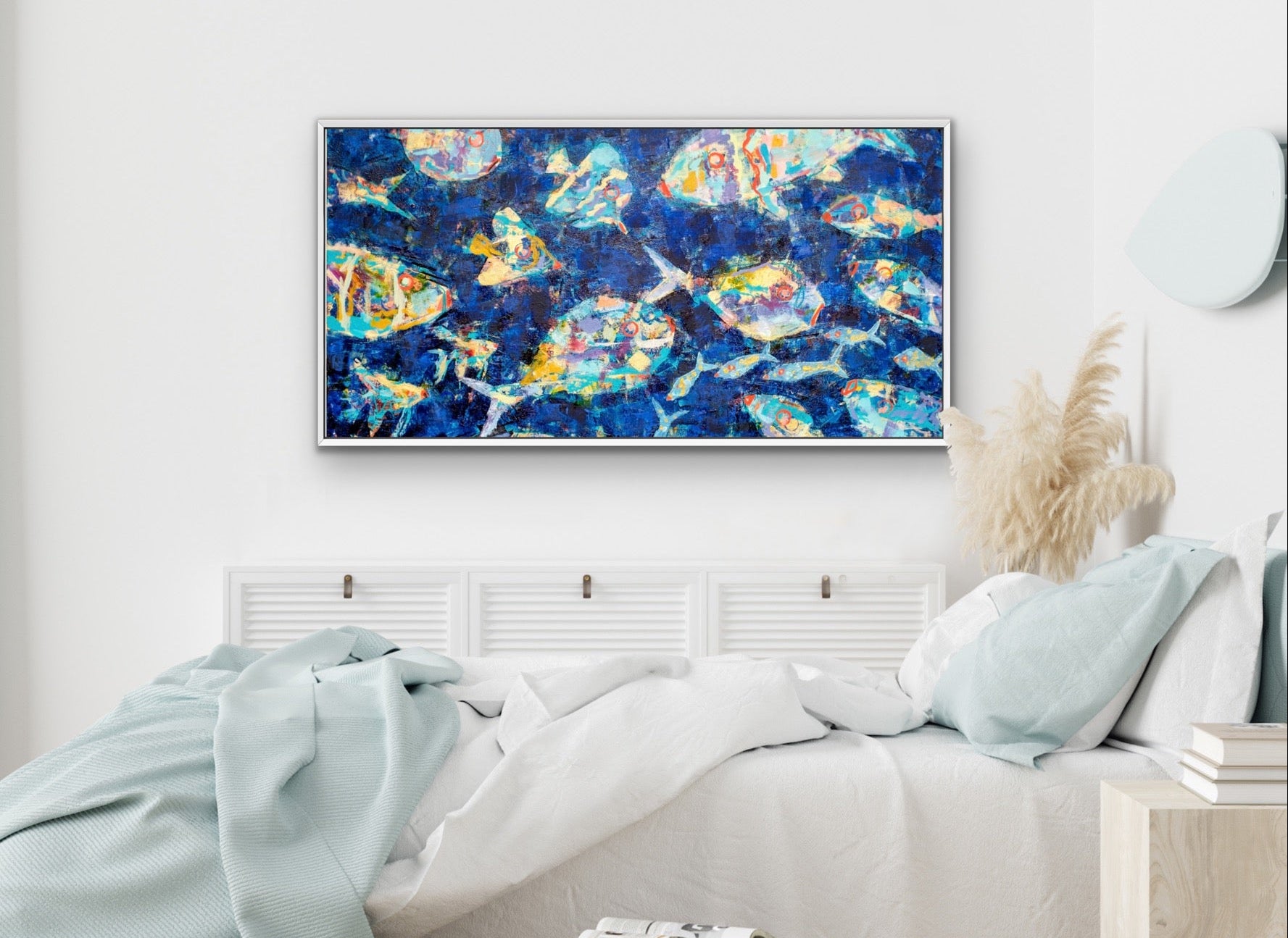 A framed abstract canvas print with a vibrant blue, gold, and white pattern, displayed on a white wall above a neatly made bed in a modern, softly lit bedroom.