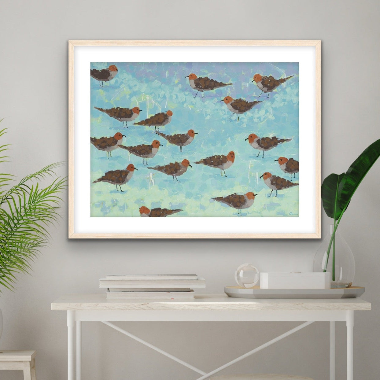 Aqua and blue background of seagulls on the beach wall art hanging above a white desk with plants and decor.
