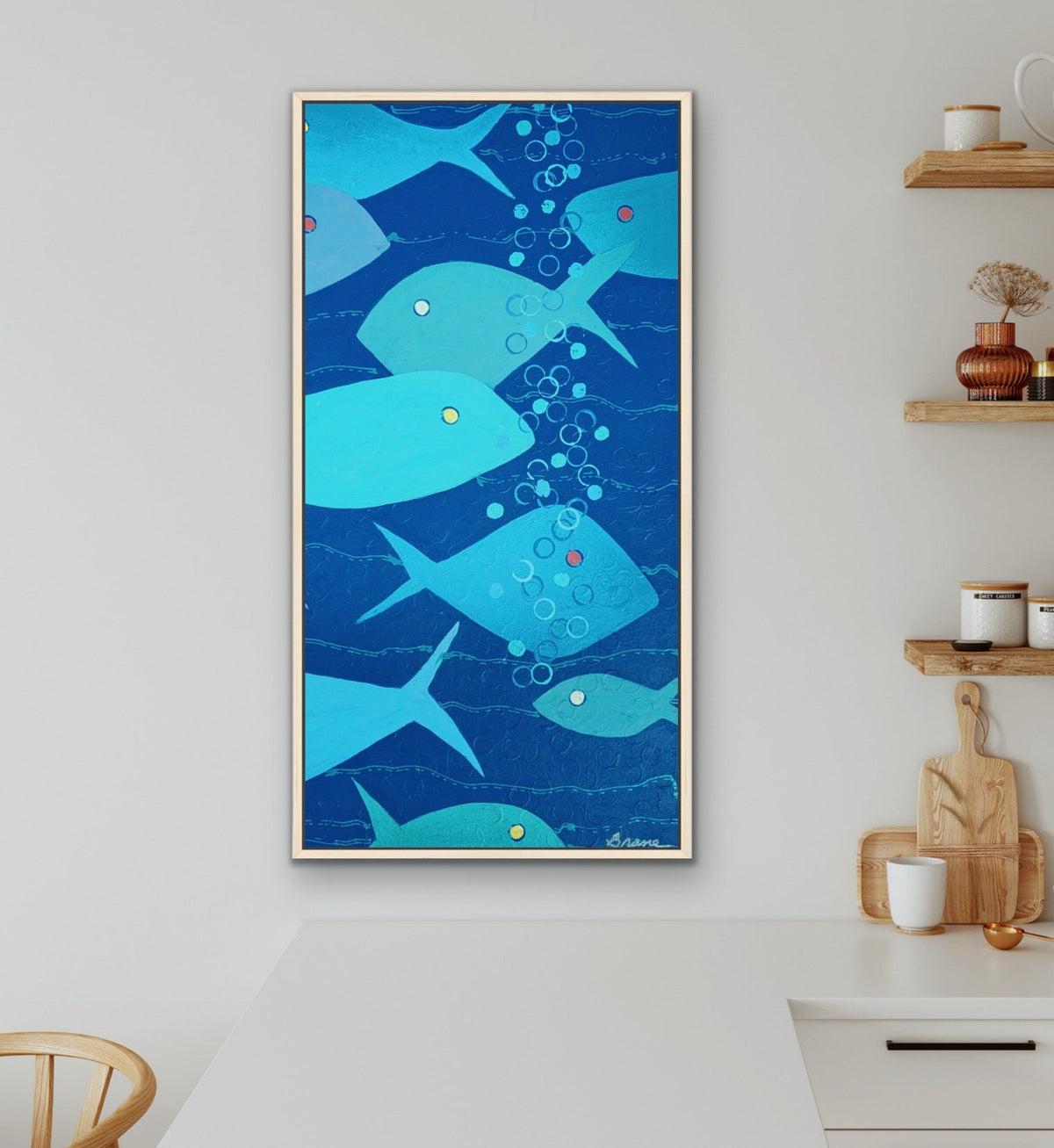 A vertical framed wall art print featuring a playful underwater scene with light blue fish and bubbles on a deep blue background, styled in a modern kitchen setting.