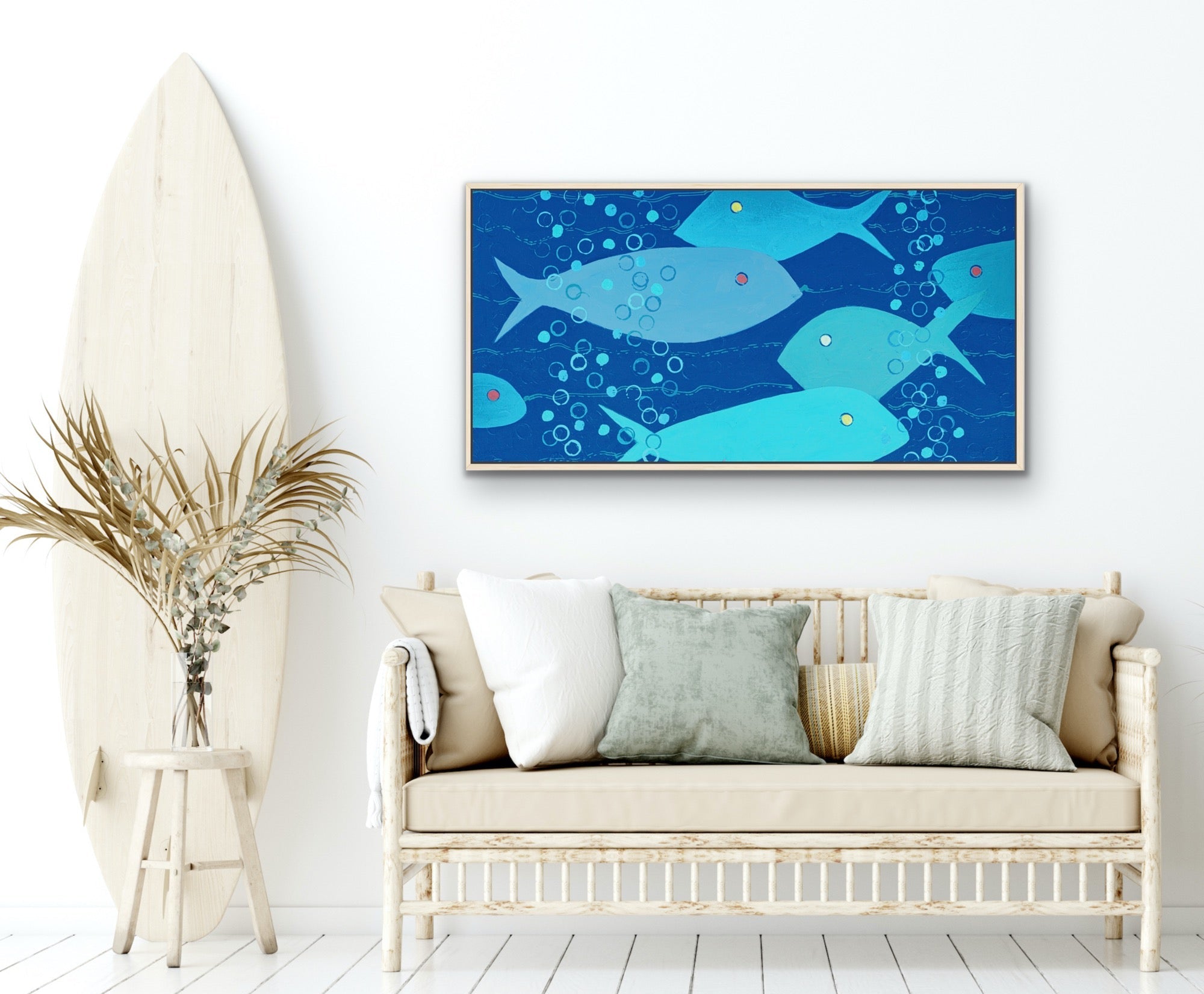 A framed horizontal canvas print featuring a playful underwater scene with light blue fish swimming in a deep blue ocean, styled on a white wall above a beige sofa with decorative pillows, next to a tall wooden surfboard.