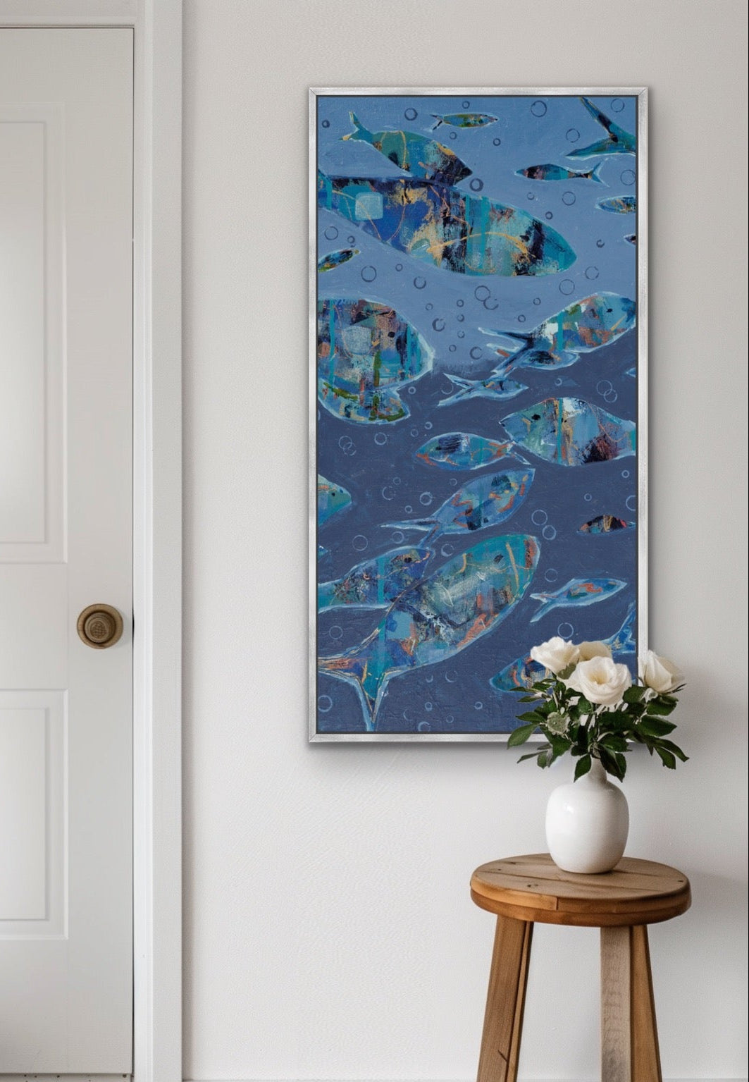 A framed vertical canvas print featuring a school of blue and teal fish, styled on a white wall above a wooden stool with a white vase of flowers.