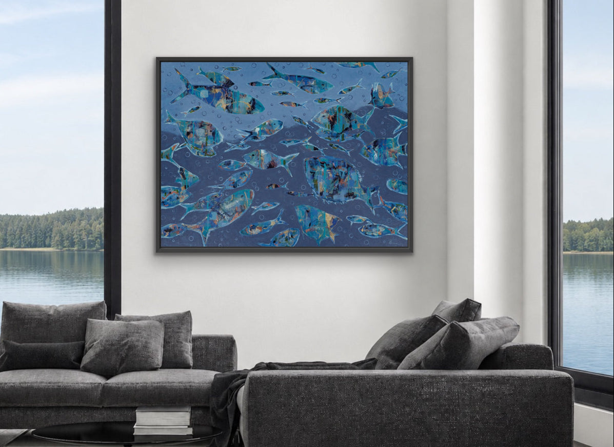 A framed abstract blue fish painting hangs on a white wall in a modern living room with two gray sofas and large windows overlooking a lake.