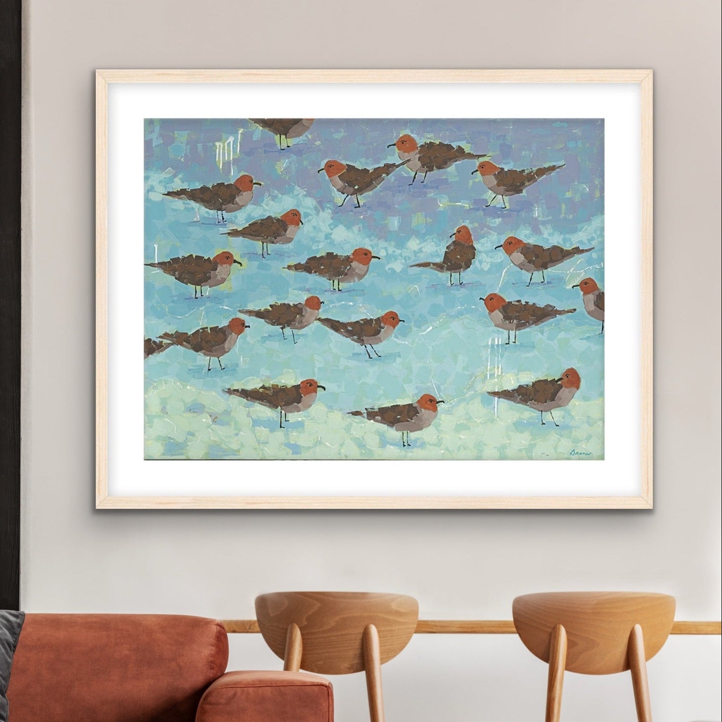 Modern coastal bird art print on cotton rag paper showing shorebirds in warm brown and coral tones in a living room setting with a brown sofa and wooden chairs.