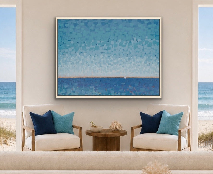 A framed horizontal art print featuring a blue and teal ocean landscape hangs on a beige wall in a bright, modern living room with two beach-view windows, white armchairs, and a wooden coffee table.
