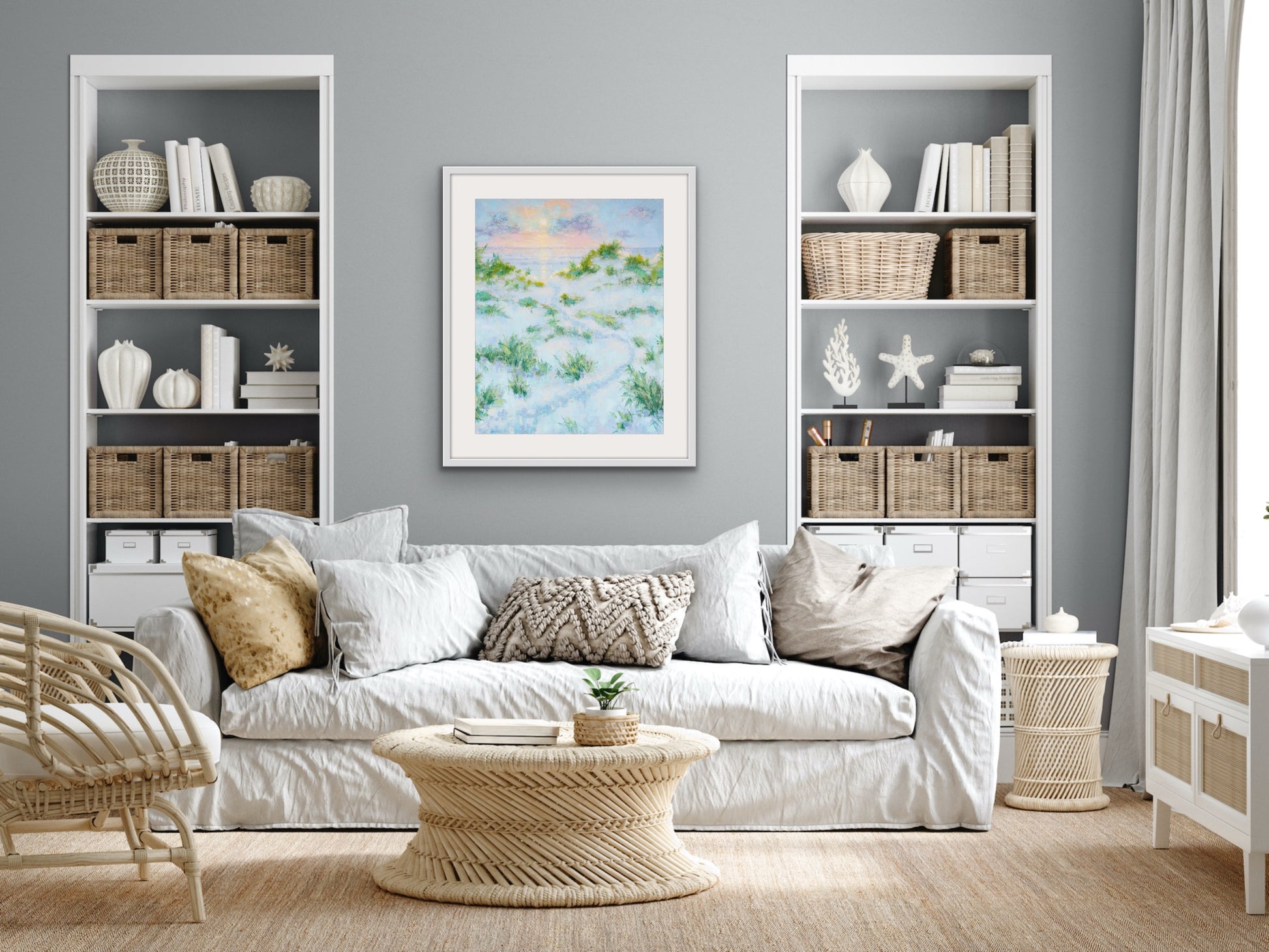 Living room with white sofa, wicker coffee table, bookshelves and a tall cotton rag coastal landscape framed with white mat on the wall.