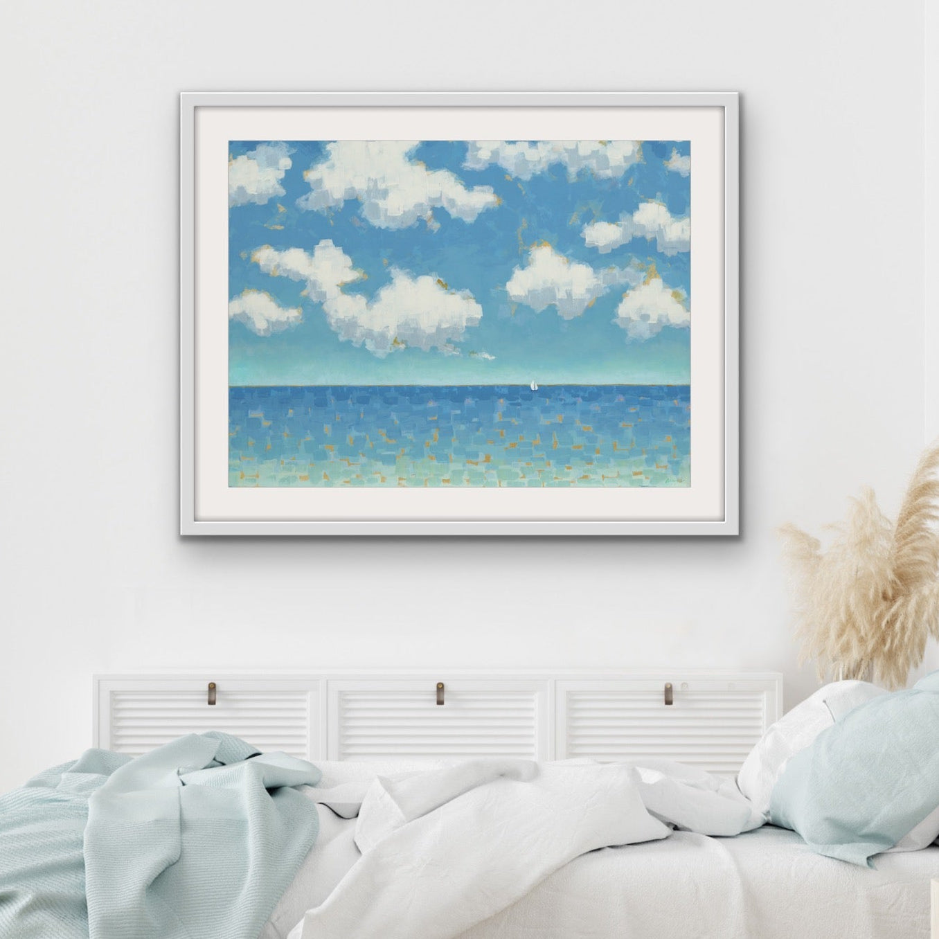 Wide horizontal ocean artwork with cloud-filled sky framed artwork of a beach scene above a bed in a bedroom.