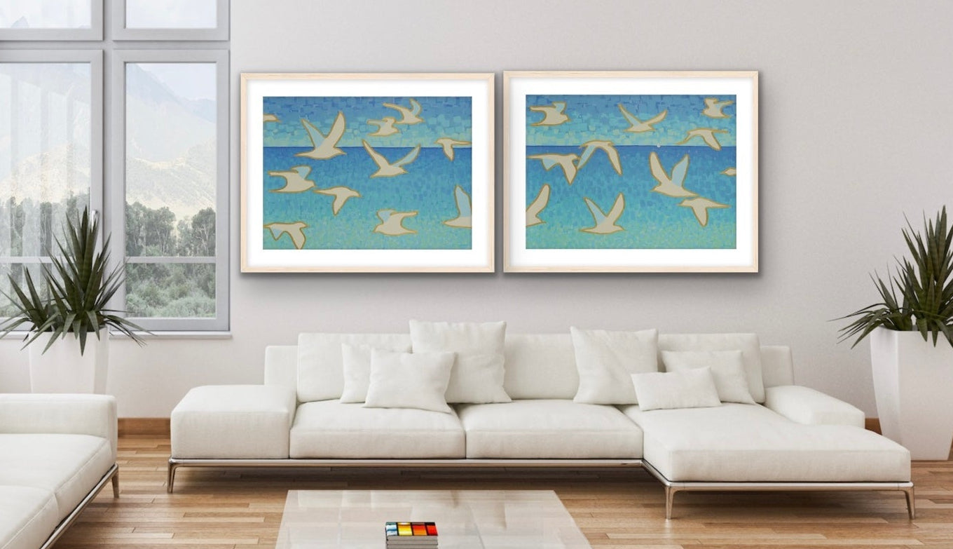 Wings of Light I & II cotton rag diptych with seagulls and sailboat on ocean horizon, modern coastal wall art by Martha Brane in a modern living room with white sofas and a glass coffee table.