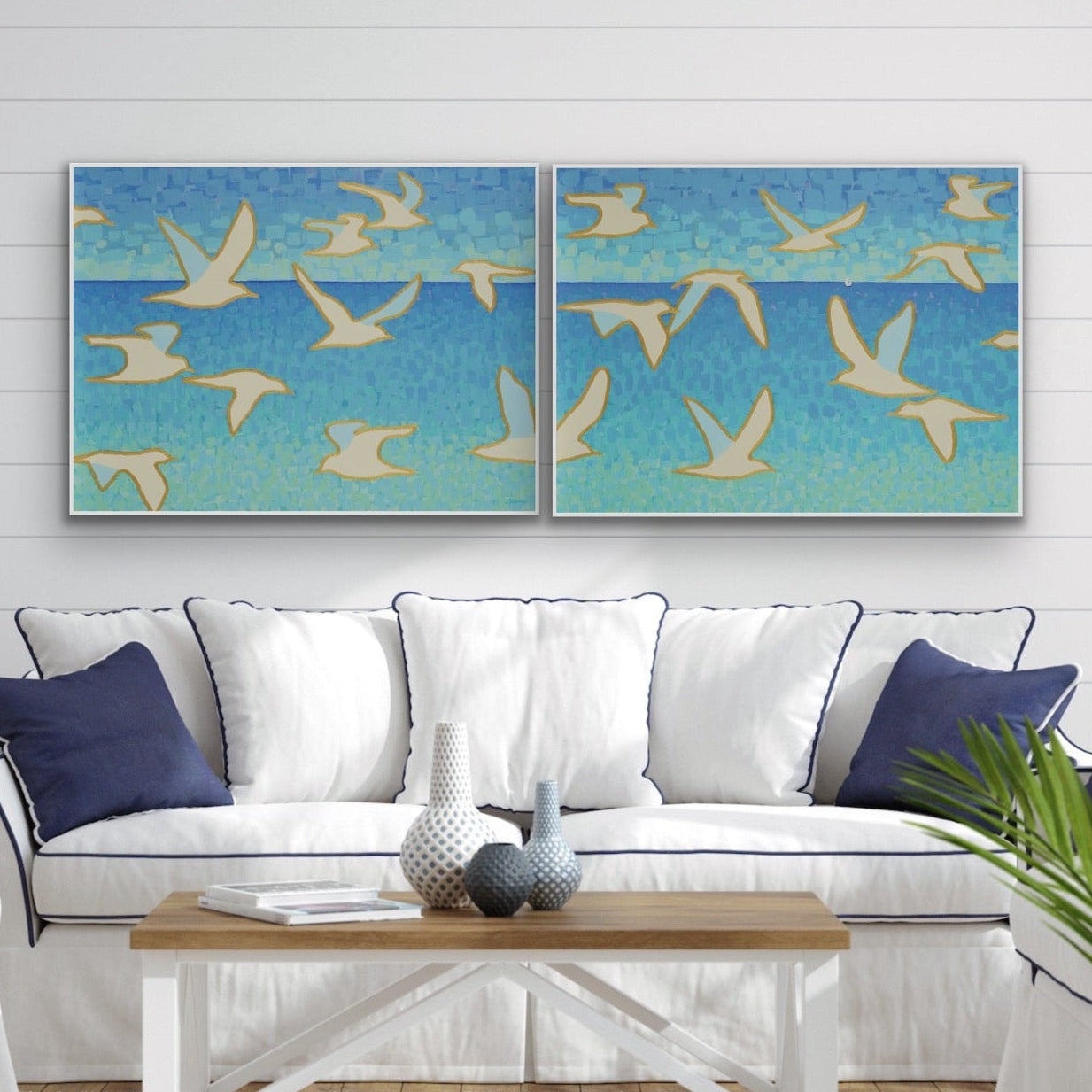 Wings of Light I & II coastal giclée canvas diptych with white seagulls and sailboat on the horizon, ocean blue wall art by Martha Brane in a living room with a white sofa, and wooden coffee table.  