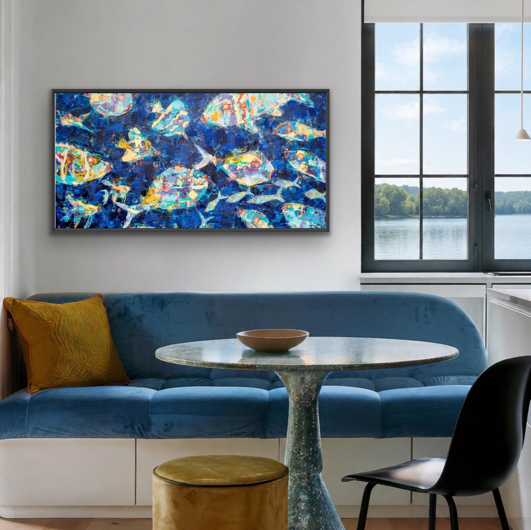 A framed abstract canvas print featuring a vibrant blue background with colorful fish, displayed on a white wall above a blue velvet sofa in a modern living room setting.