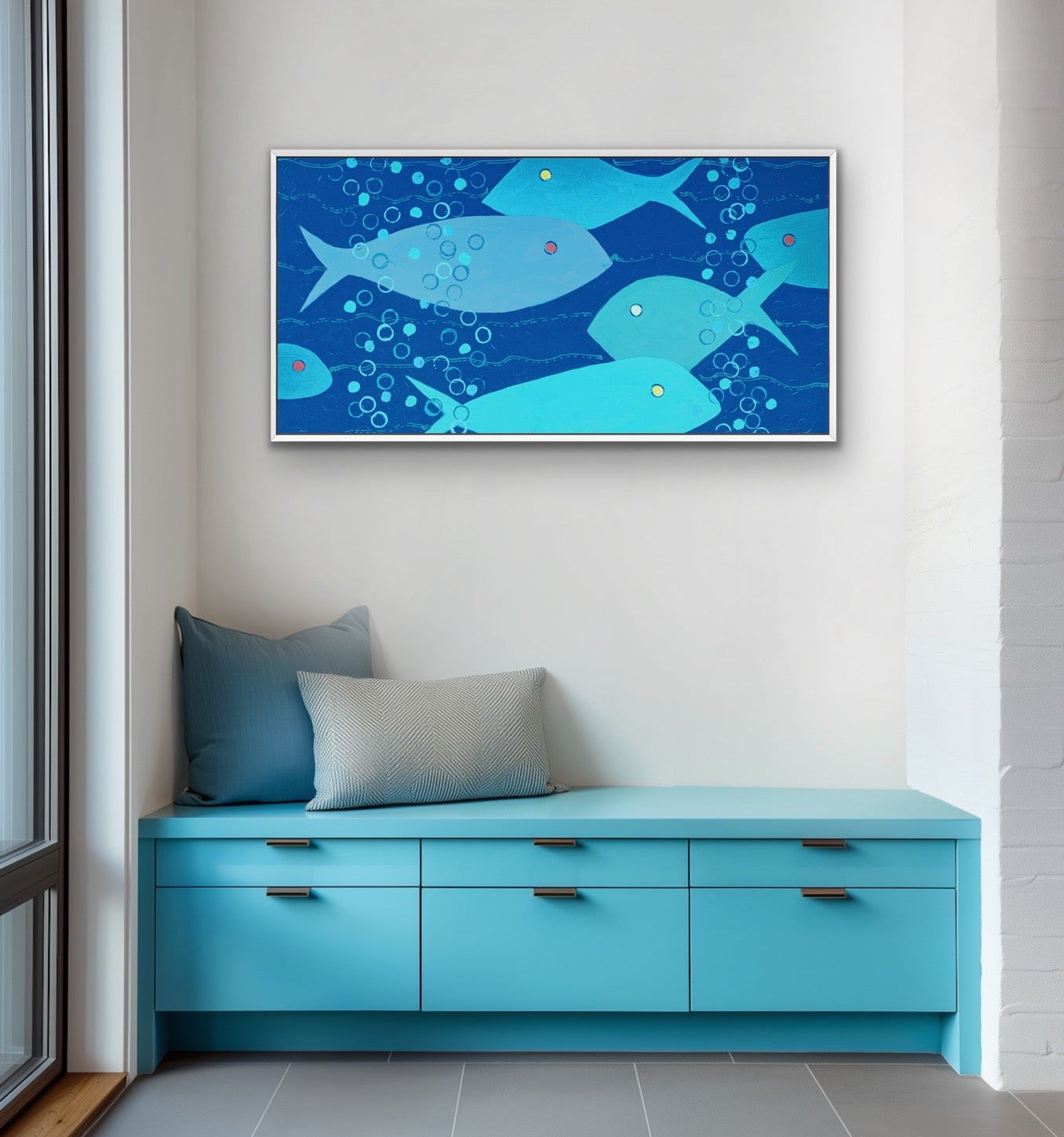 A framed horizontal art print depicting blue and light blue fish swimming underwater with bubbles, displayed on a white wall above a bright turquoise bench with pillows.