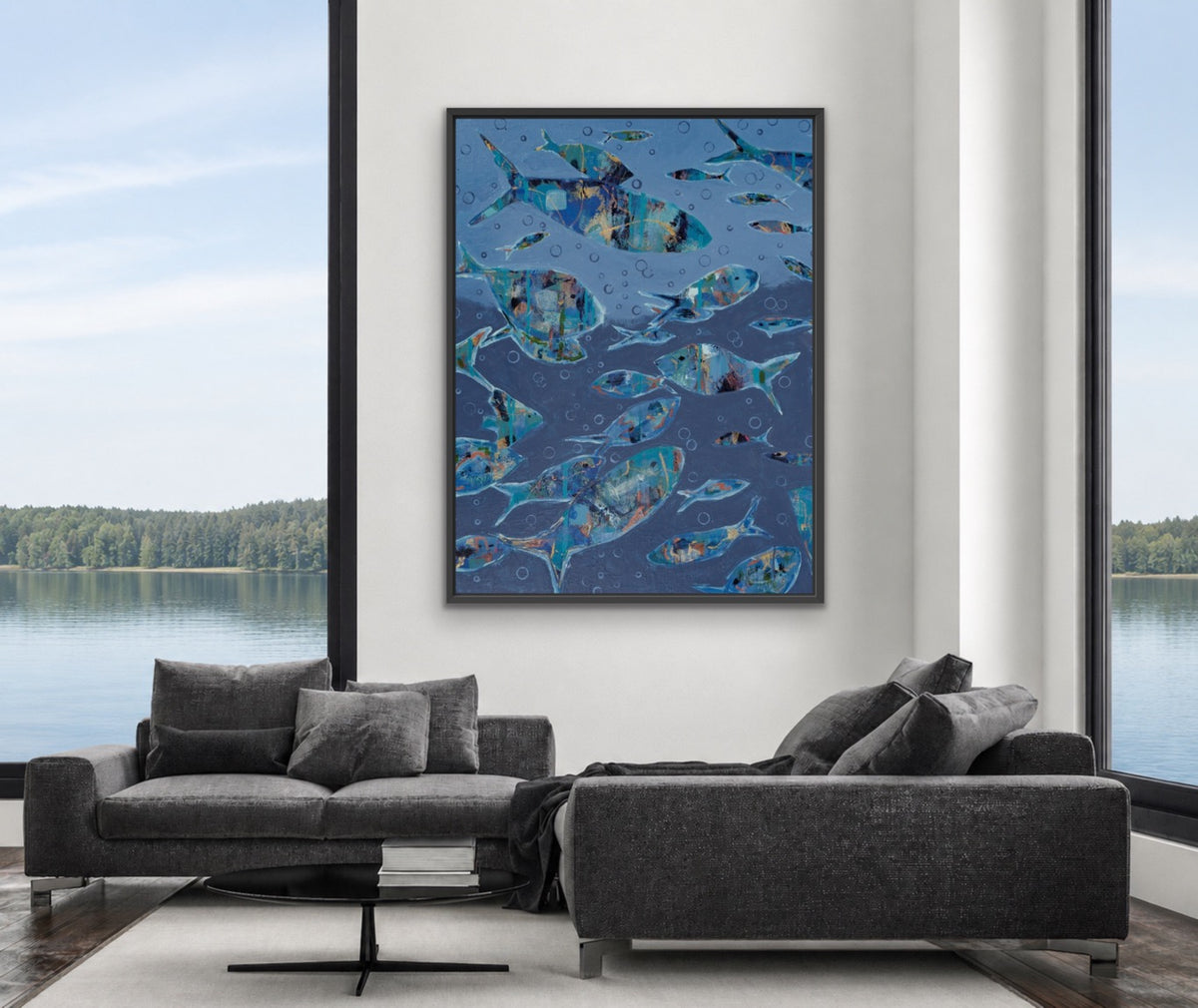 A framed vertical art print depicting a school of blue fish on a dark blue background, displayed on a white wall in a modern living room setting with a gray sectional sofa and large windows overlooking a lake.