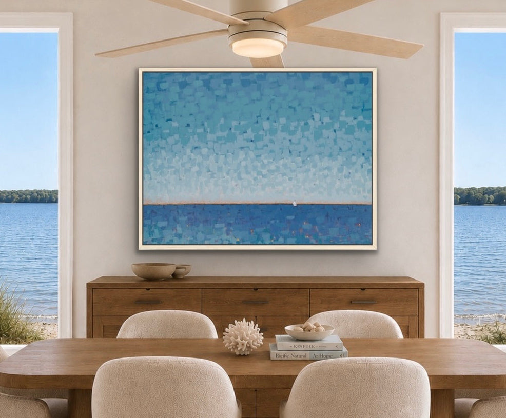 A large square framed art print depicting a blue textured sky and ocean, displayed on a beige wall in a modern dining room setting with a wooden table and chairs.