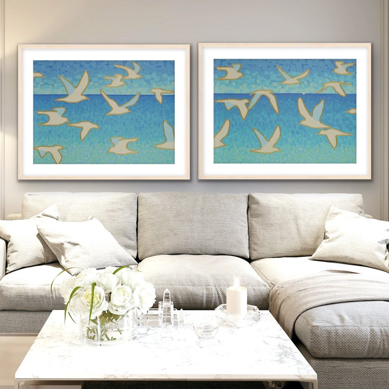 Wings of Light I and II modern coastal diptych, ocean horizon with white birds and sailboat, archival cotton rag prints in a modern living room with a gray sectional sofa, and a coffee table.