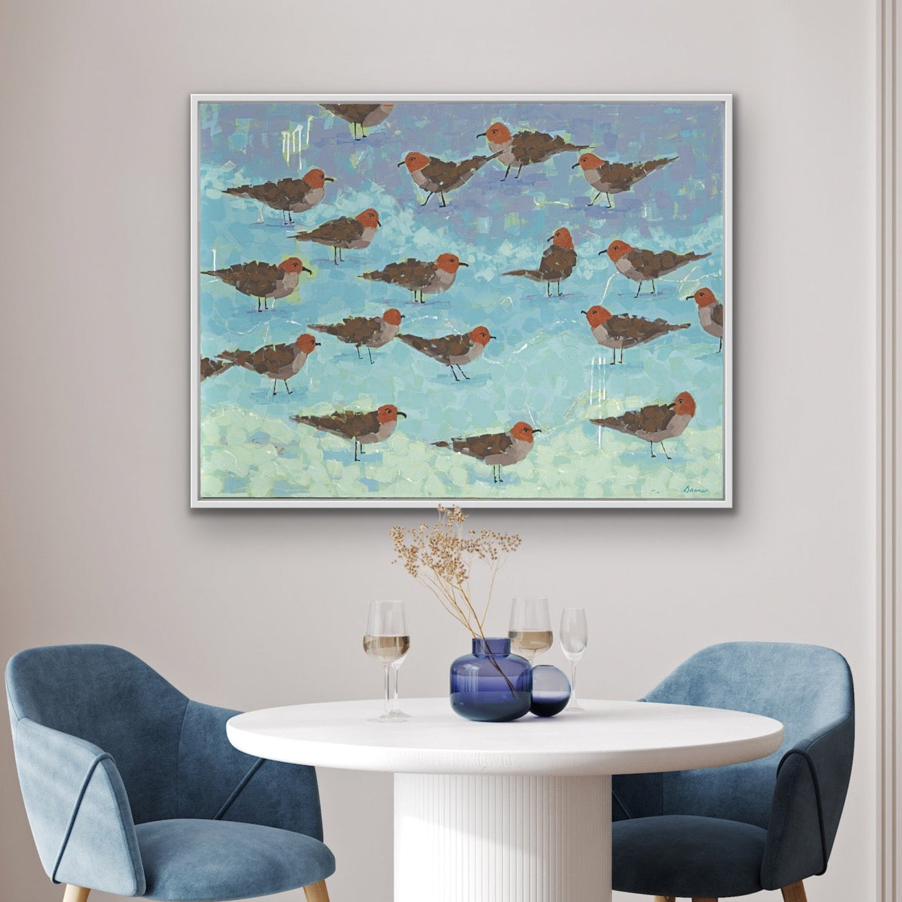 Contemporary coastal bird art “Morning Briefing II” framed in white wood above dining area with a round table and blue chairs.