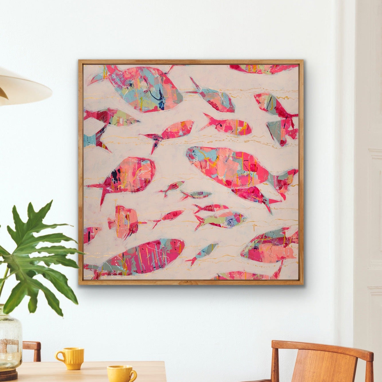 Modern coastal artwork with colorful fish and gold accents on a square canvas on a wall above a dining table with chairs.