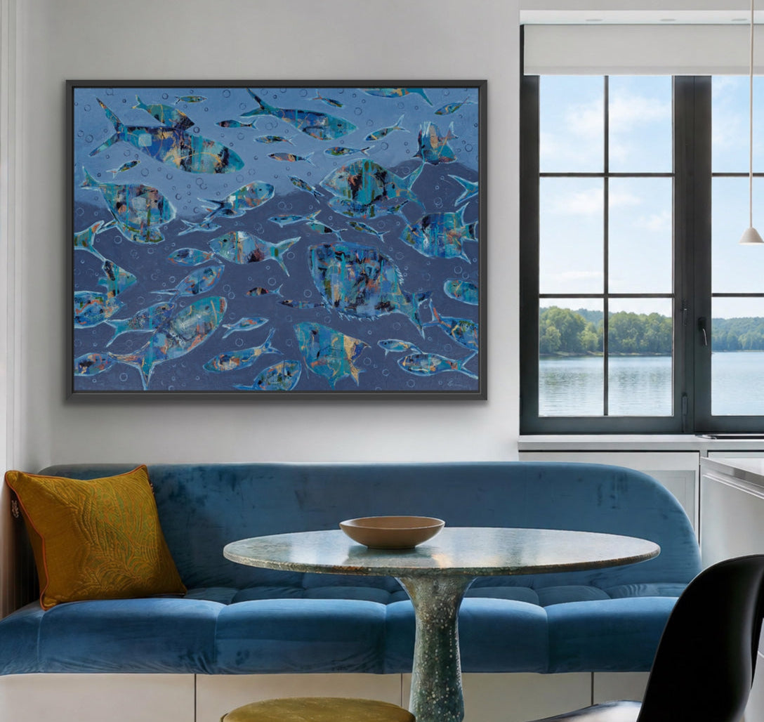 A large framed abstract canvas print featuring blue and teal fish patterns hangs on a white wall in a modern living space with a blue velvet sofa, a round marble-top table, and a black chair.