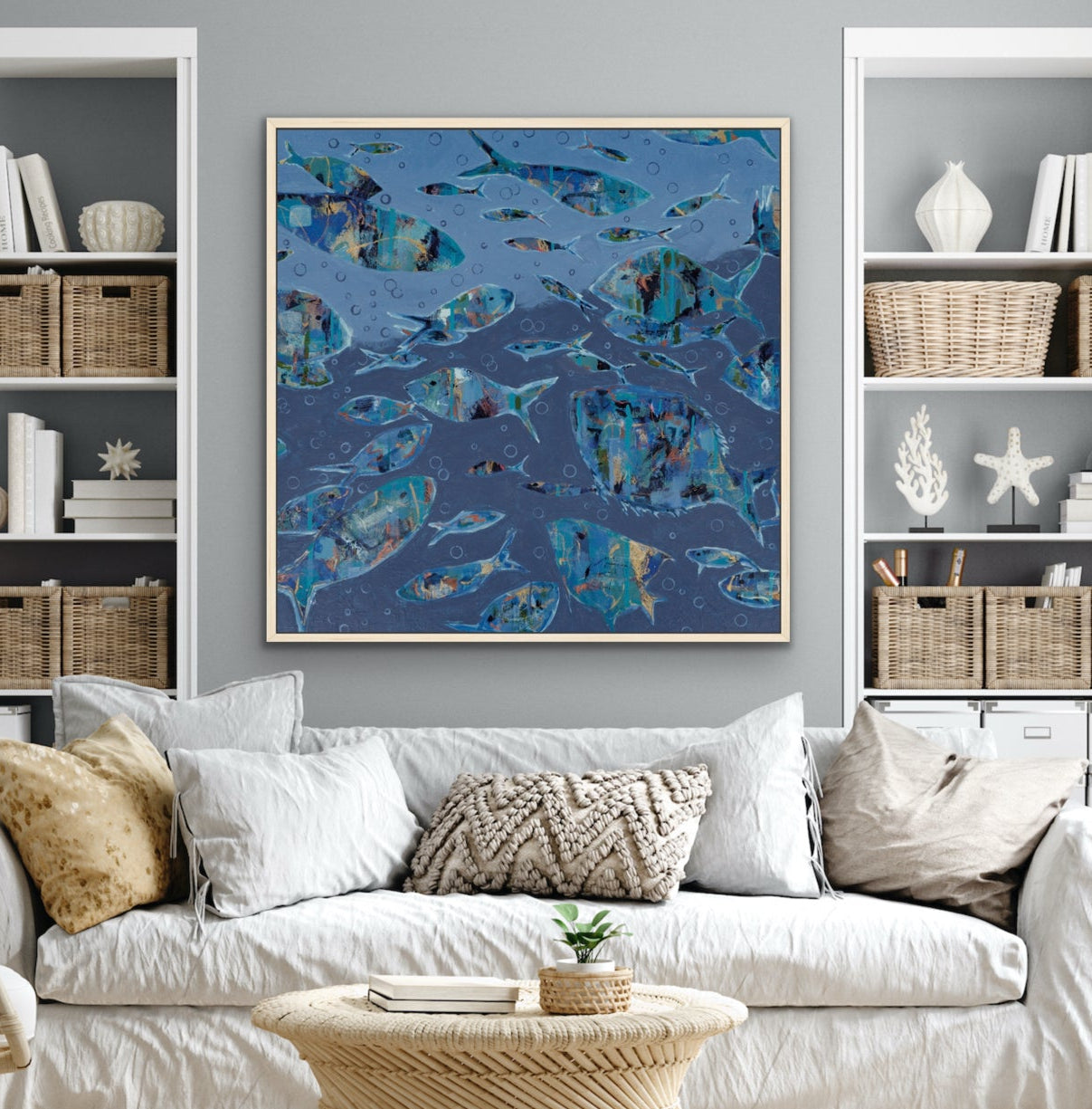 A framed abstract blue and teal abstract fish painting hangs on a gray wall above a white sofa in a styled living room with shelving, a rope coffee table, and neutral decor.