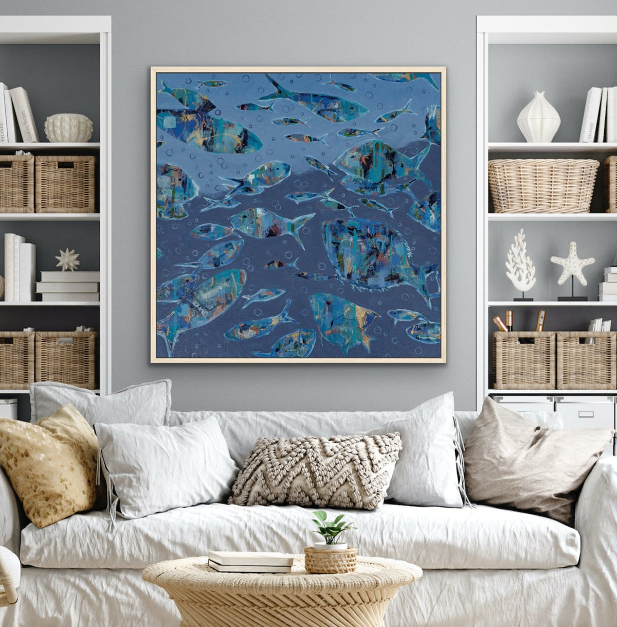 A framed abstract blue and teal abstract fish painting hangs on a gray wall above a white sofa in a styled living room with shelving, a rope coffee table, and neutral decor.