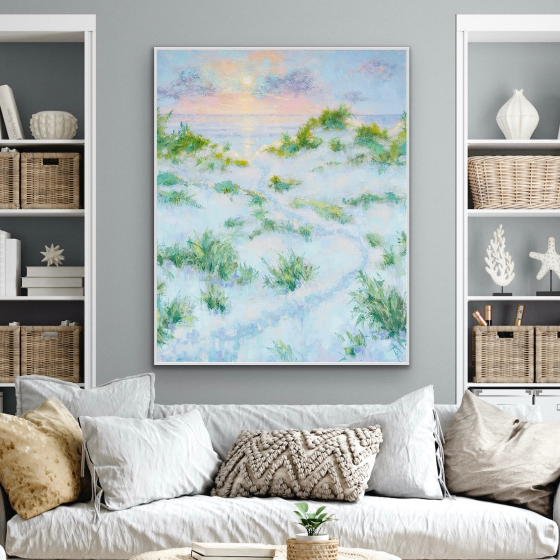 Living room with a statement sunrise seascape with dunes painting on the wall, white sofa, and bookshelves.